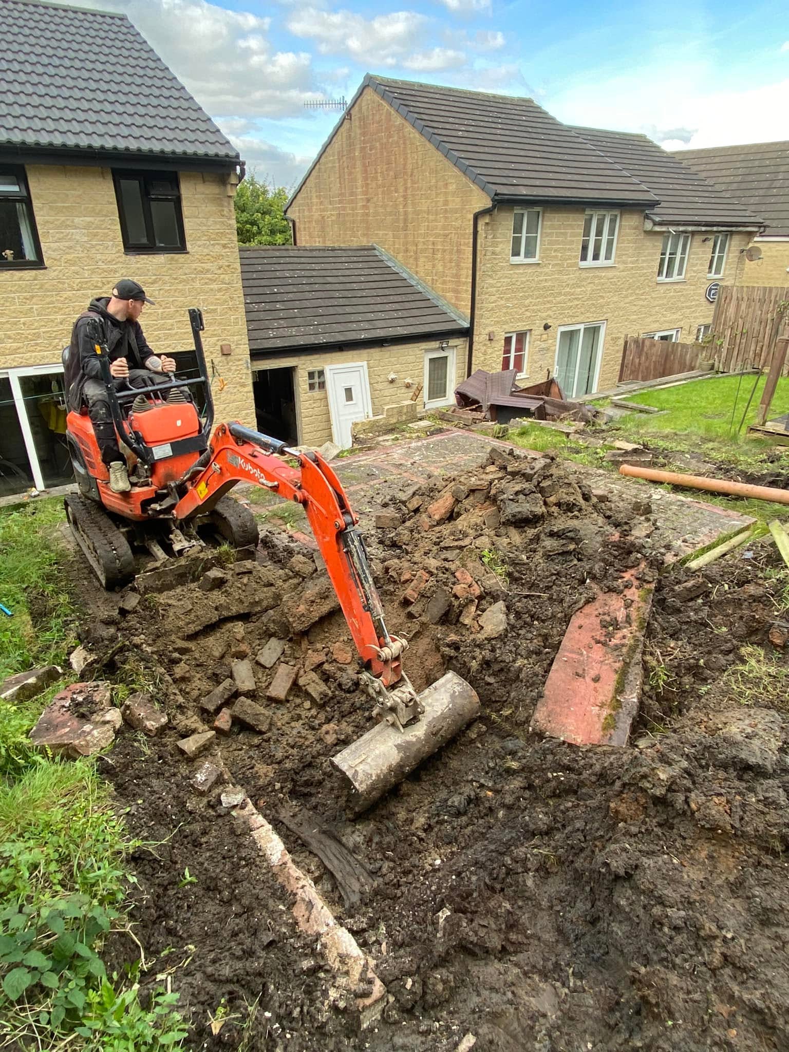 A small red excavator is digging up dirt and rubble in a residential backyard, with two brick houses in the background under a partly cloudy sky.