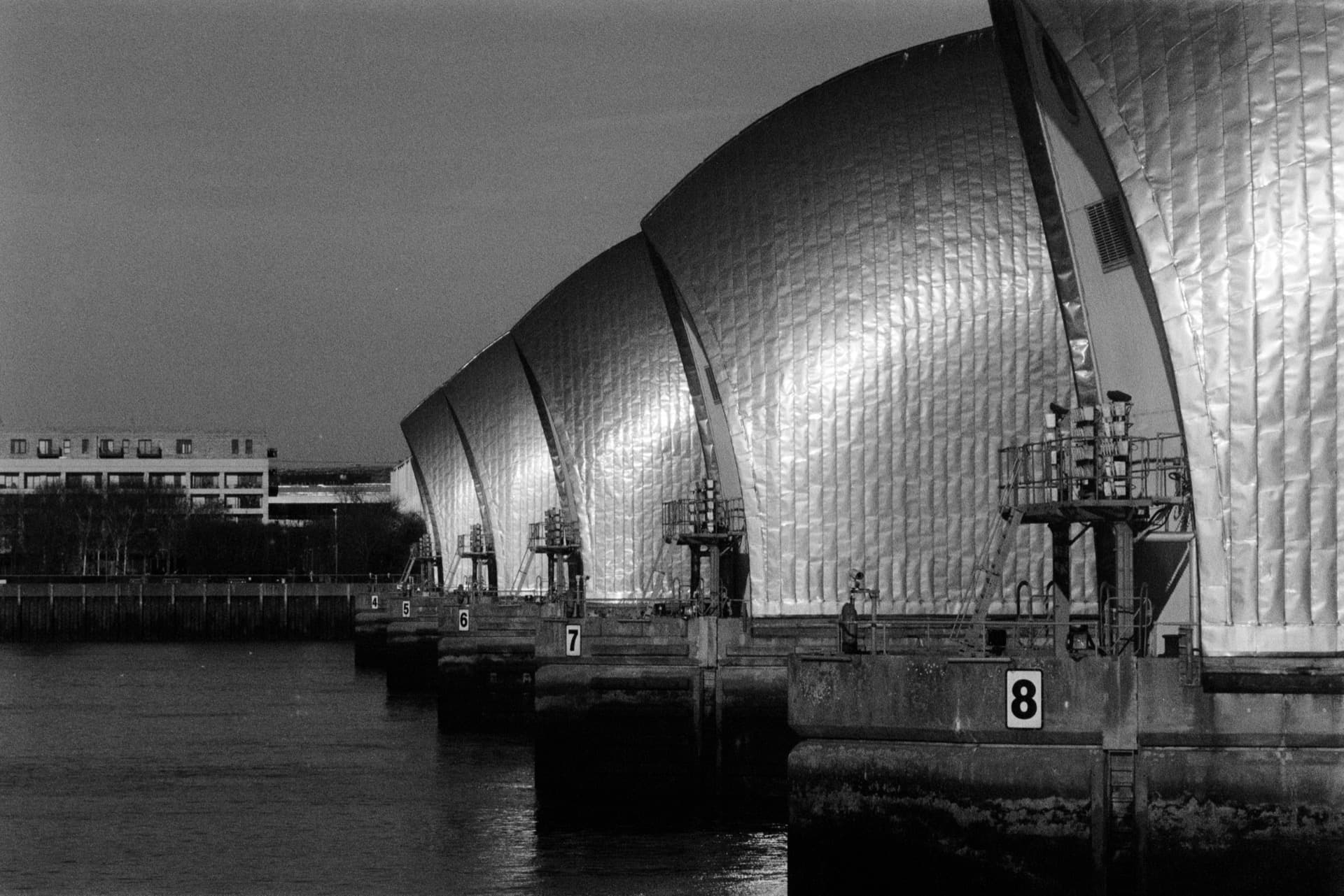 Thames Barrier with illuminated curved steel hoods at dusk, numbered pier structures (6-8) in foreground, modern residential development visible beyond