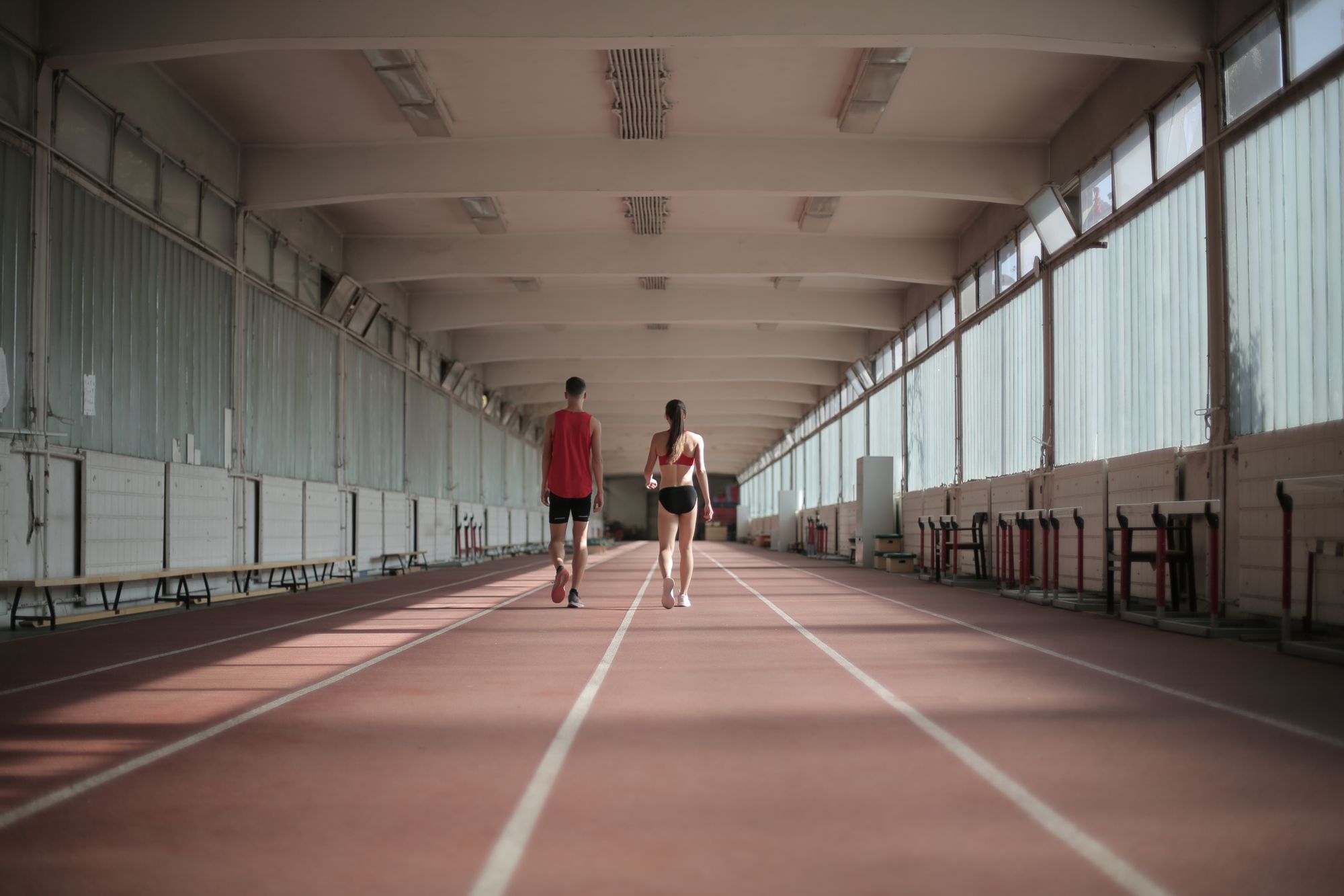 Back view of a man and woman side by side on an indoor running track, walking down the lane together under a long covered corridor.