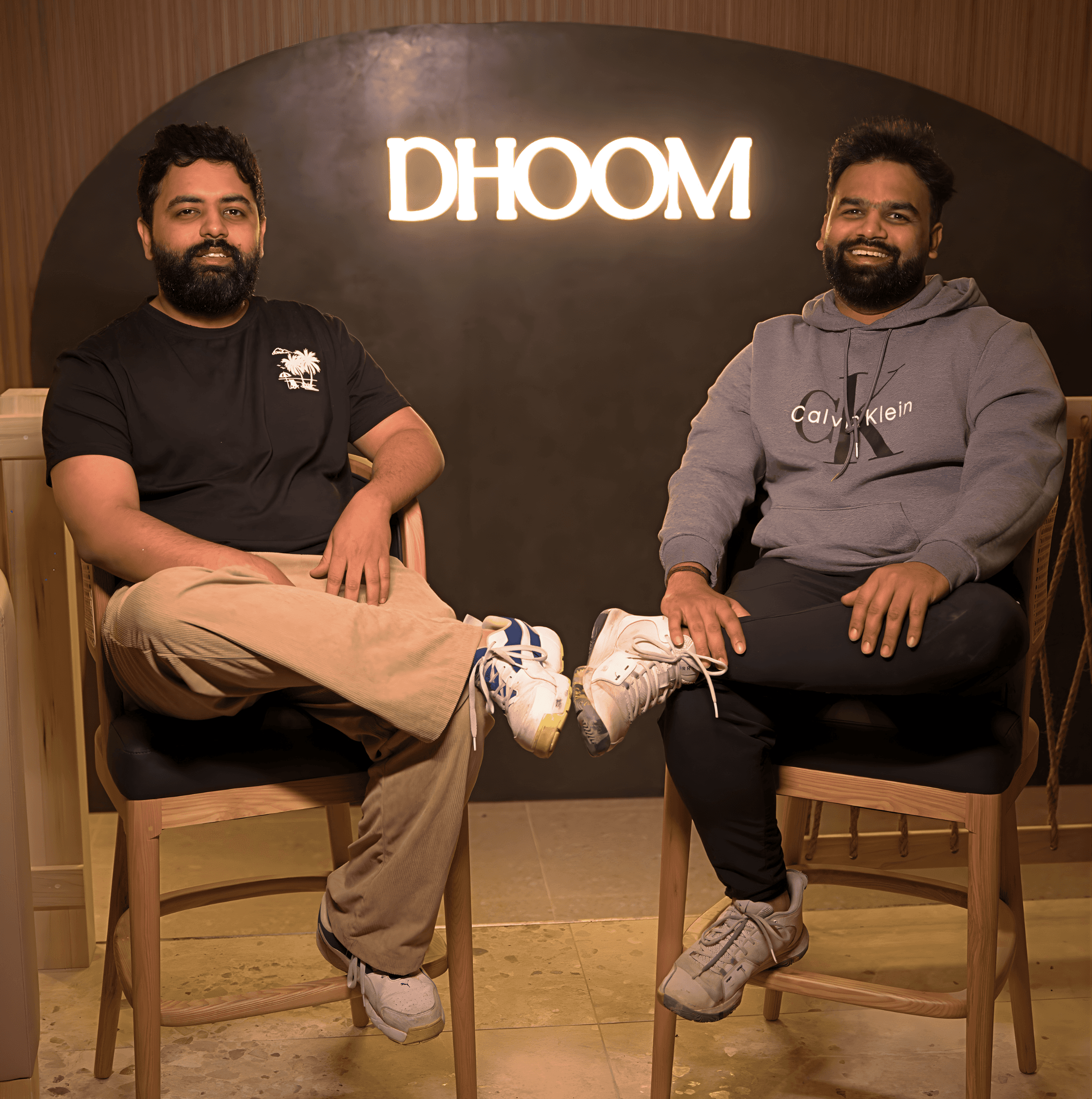 Founders Sajid Rayani and Sohail Rayani seated inside Dhoom Toronto, standing in front of the illuminated Dhoom logo, representing the vision and leadership behind the modern Indian fine dining restaurant.