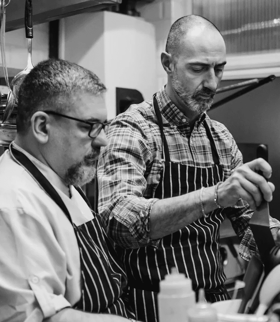 Two chefs working side by side in a kitchen, both wearing striped aprons. One chef in glasses focuses on a task in front of him, while the other, in a colourful checked shirt, concentrates as he uses a kitchen tool. Cooking equipment and bottles are visible in the foreground.