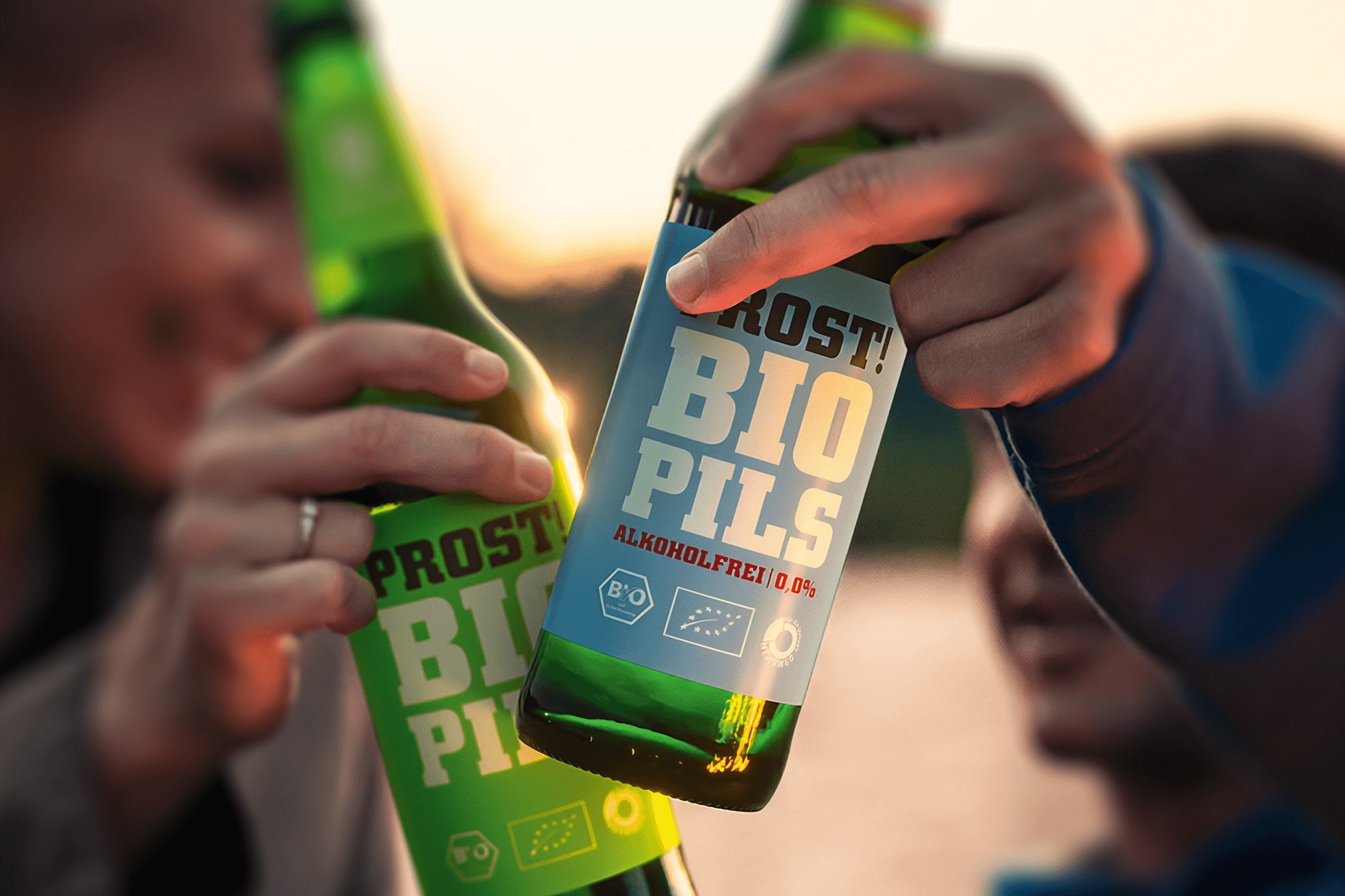 Two hands hold up green beer bottles against a sunset.