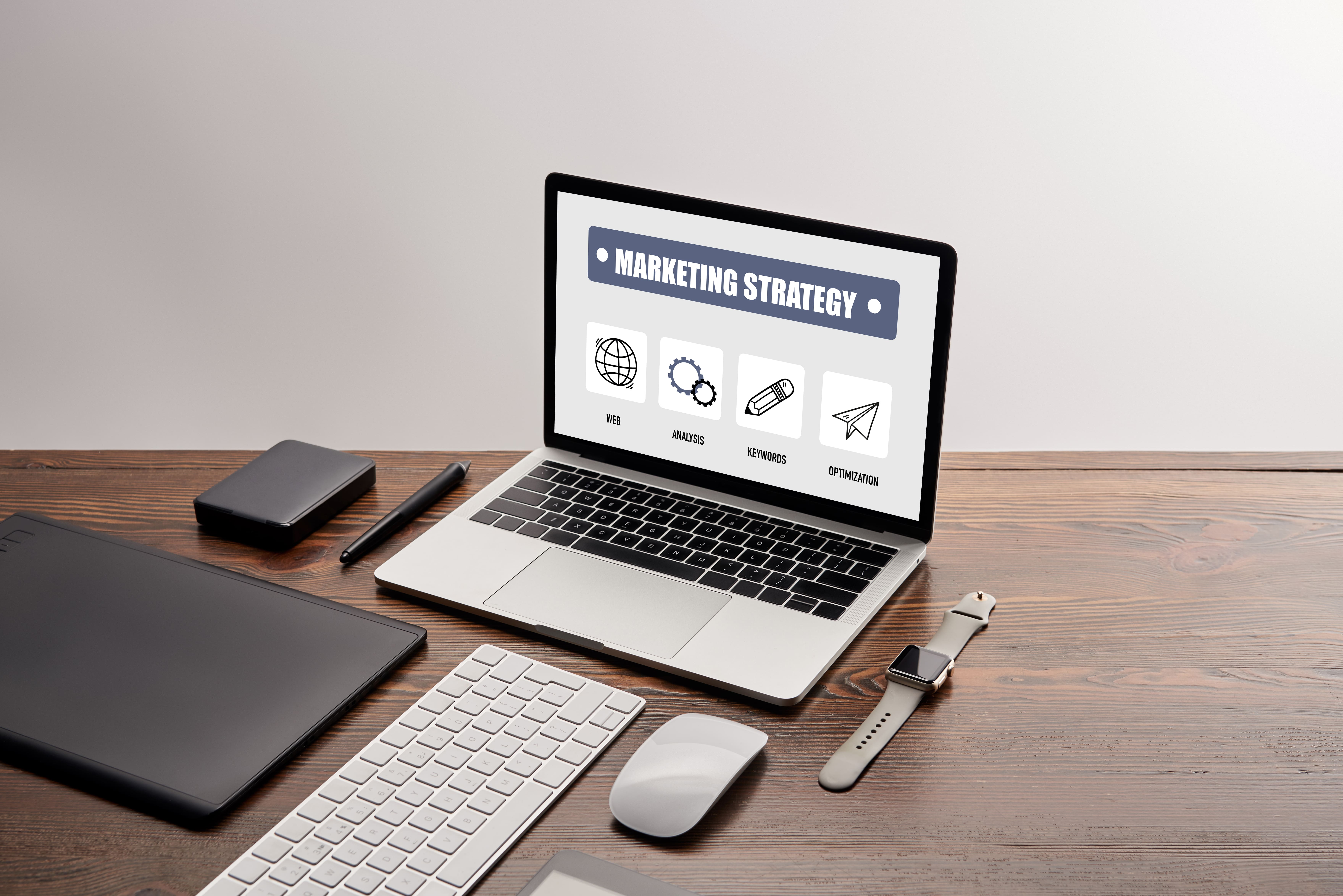 Flat lay of a laptop screen displaying "Marketing Strategy" with icons for web, analysis, keywords, and optimization, surrounded by digital gadgets like a smartwatch, stylus, and wireless keyboard on a wooden desk — representing digital marketing planning and online strategy workspace.