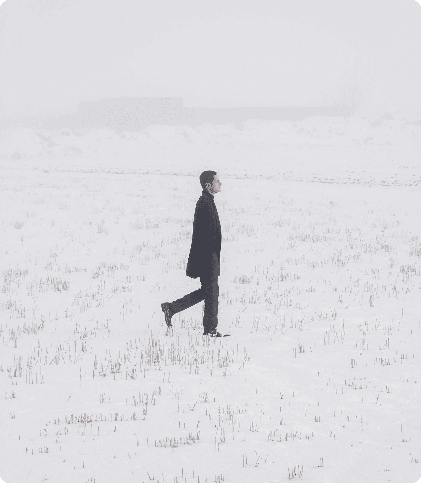A person in a long black coat walks across a snow-covered field on a foggy winter day. Sparse grass pokes through the snow, while the distant landscape fades into thick mist, creating a quiet, minimalist atmosphere.