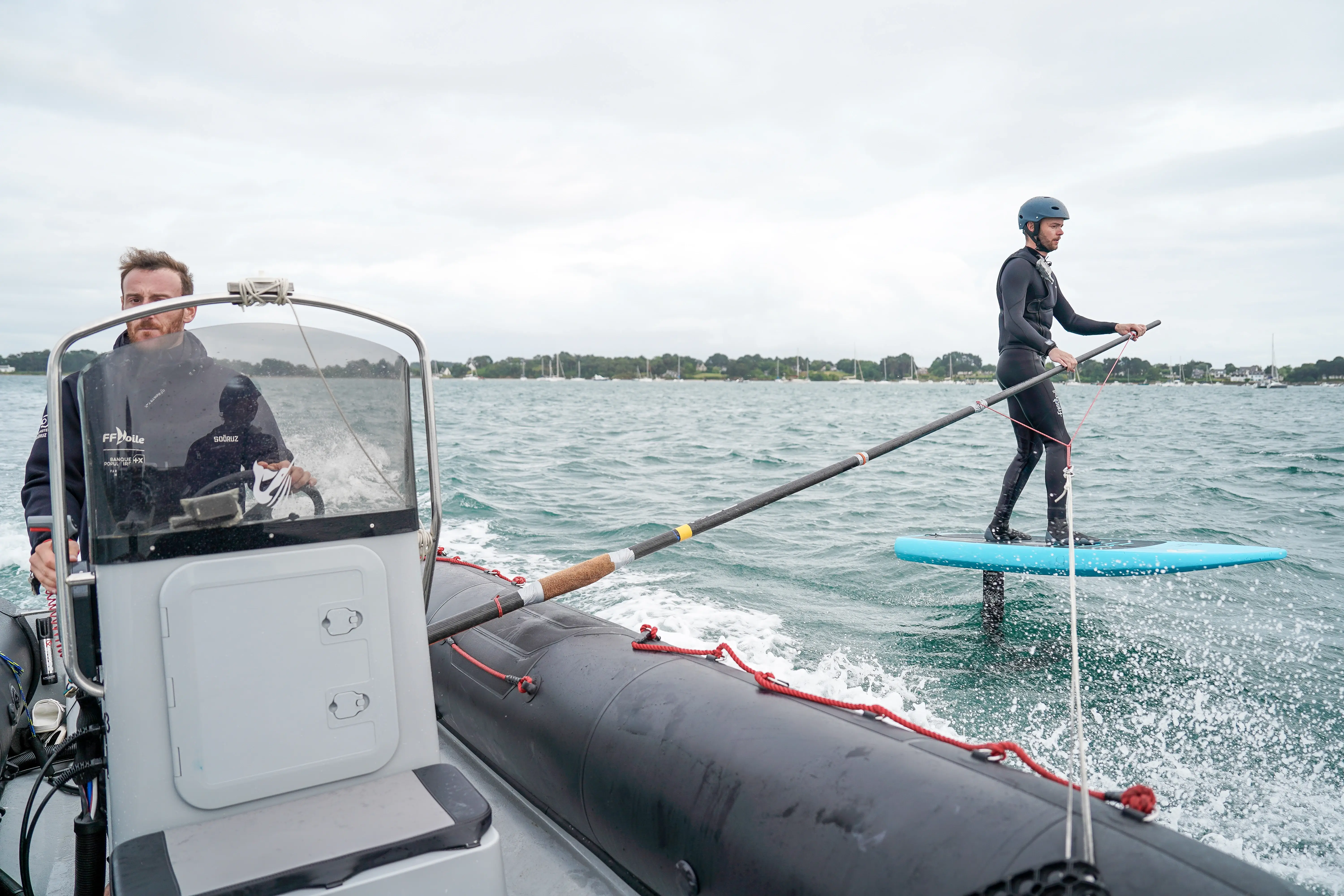 Une personne sur planche à foil, vol au dessus de l'eau et se fait tracter par un bateau en se tenant à une barre latéral