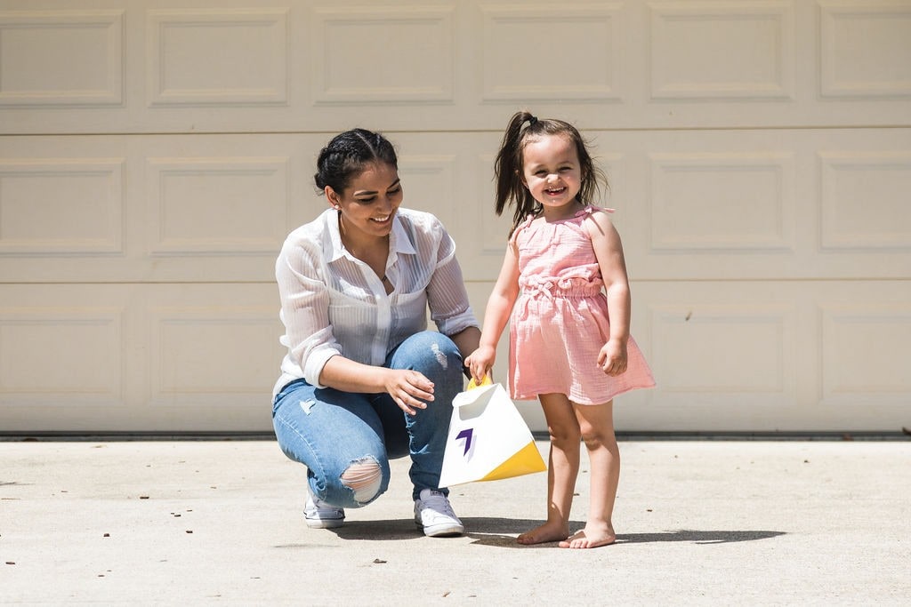 A woman crouches on a driveway in front of a garage door, smiling at a young girl in a peach dress holding a delivery package.