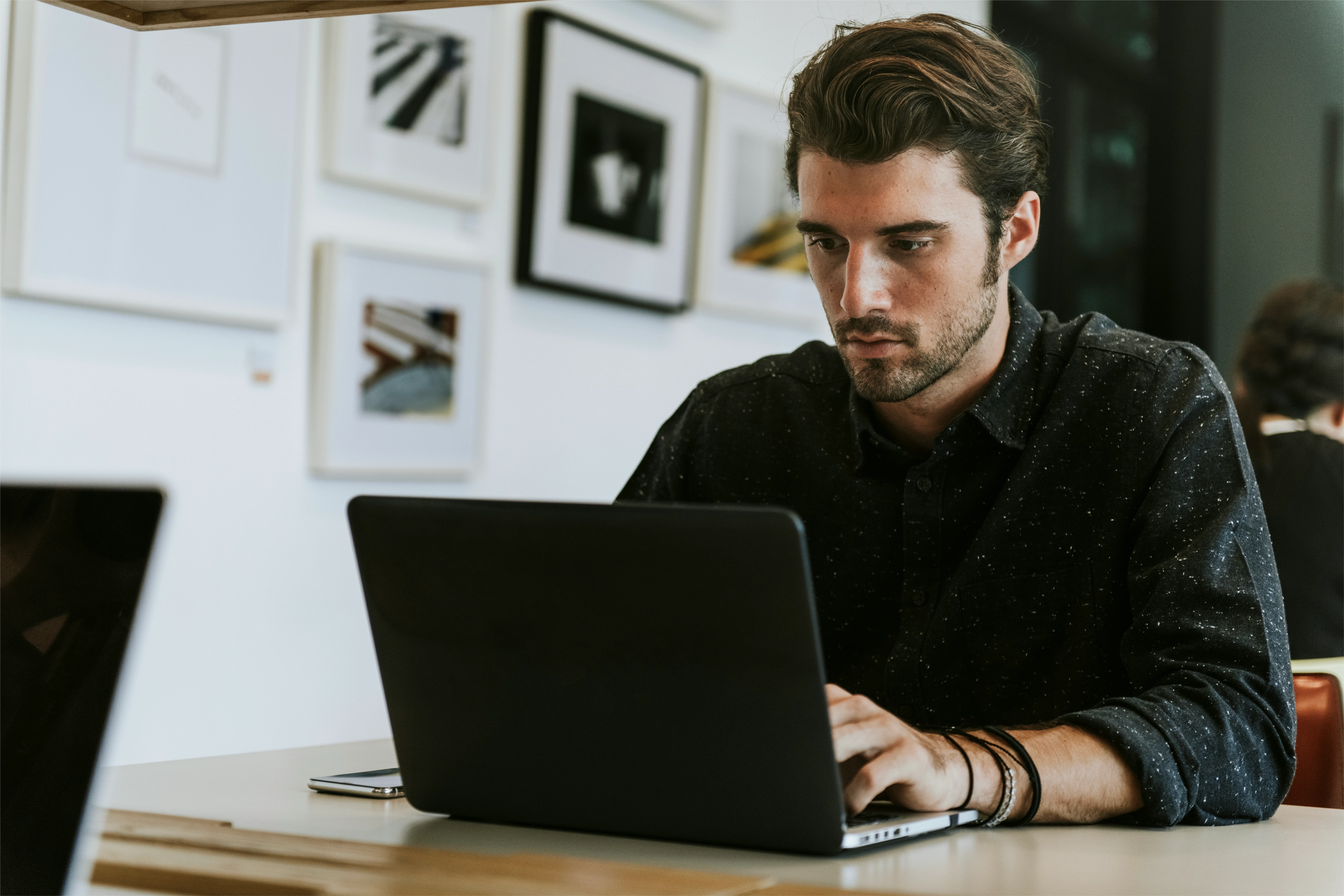 A man in a black shirt focused on his laptop, working at a table with light-colored wood.