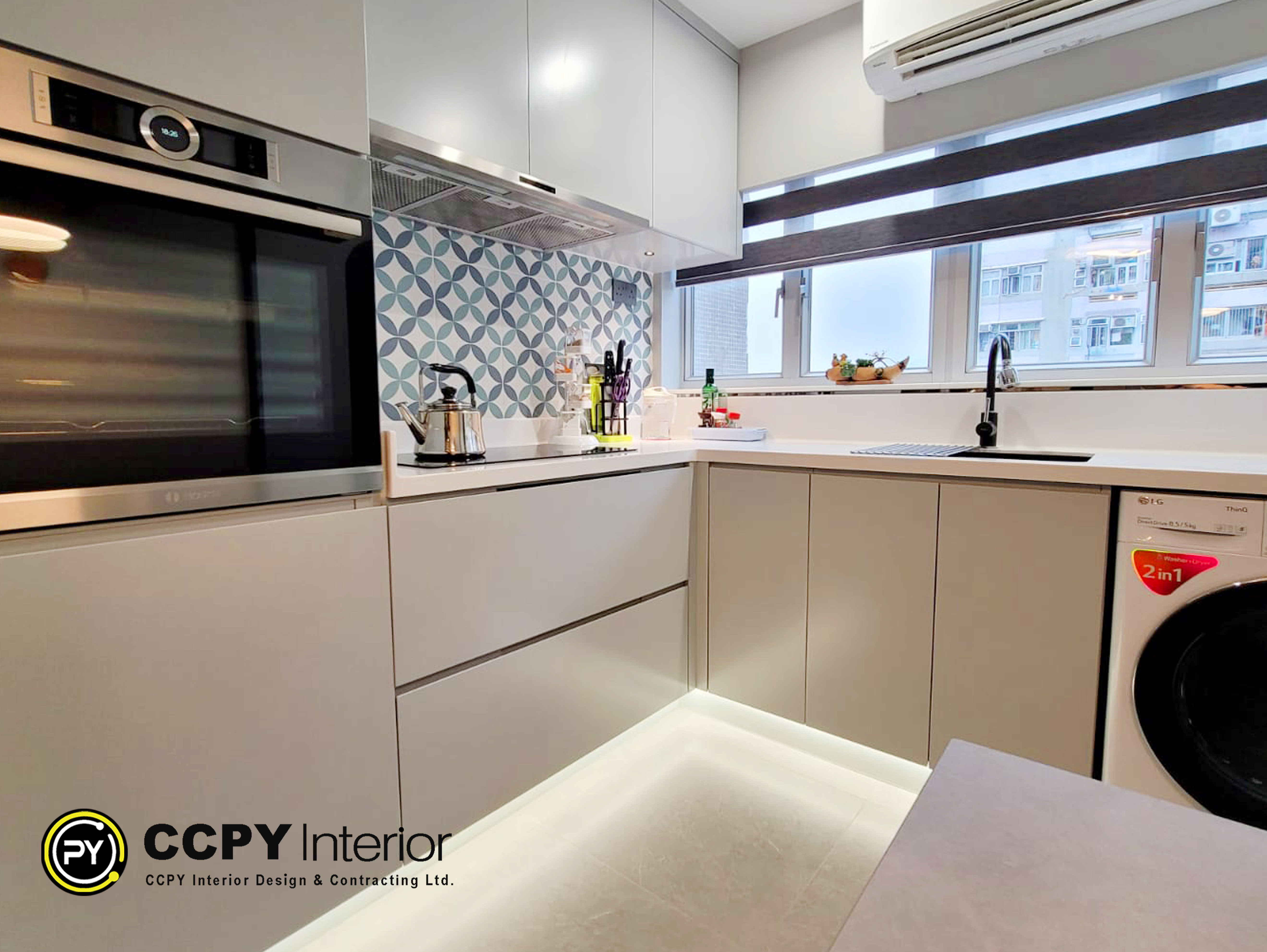 This image shows a modern kitchen interior designed by CCPY Interior Design & Contracting Ltd.Color Palette: The kitchen features light grey cabinets with integrated handles and a white countertop.Backsplash: A patterned backsplash with blue and white circular designs adds a decorative element behind the cooktop.Appliances: The design includes a built-in oven, a stainless steel kettle, and a compact washer/dryer unit.Lighting: Under-cabinet lighting is installed to illuminate the countertop area