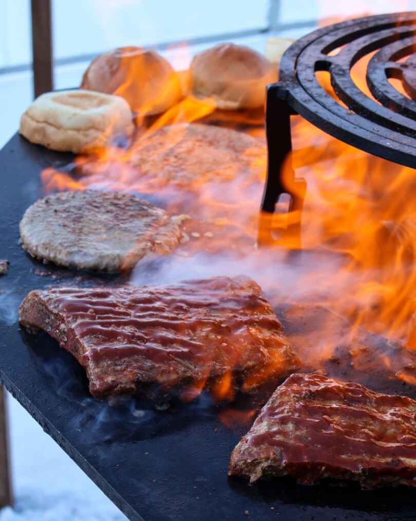 Meat patties grilling over open flames on a flat-top grill with buns warming in the background.