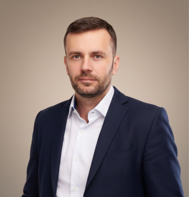 A confident person with short, styled hair and a leather jacket poses against a dark backdrop.