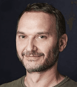 Close-up portrait of a freckled woman with soft pink hair and star earring, natural light, white tank top.