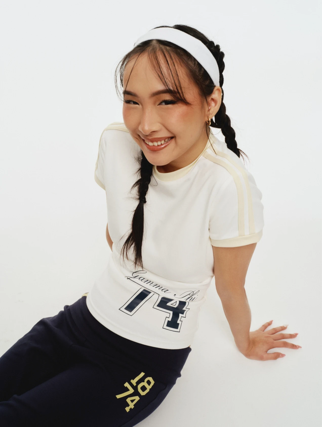Smiling model seated on white backdrop wearing a cream ringer tee with “Gamma Phi 74” and navy leggings with “1874,” plus a white headband and braided hair.