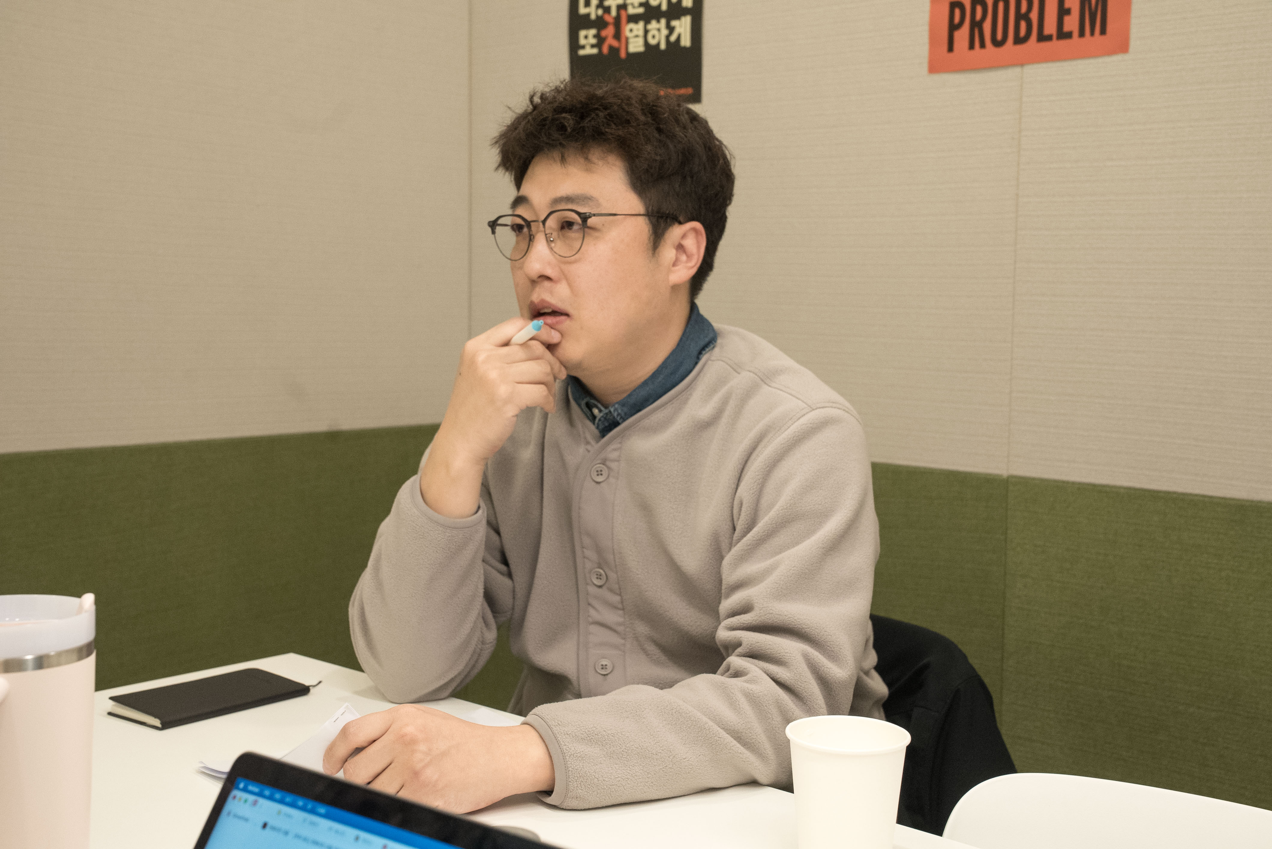 노트북과 종이가 놓인 테이블에 앉아 손에 펜을 들고 생각하며 이야기하는 어지환 과장의 모습입니다.