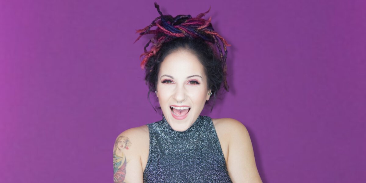 A woman with vibrant multicolored dreadlocks and a glittery top smiles joyfully against a purple background. Her expression is lively and cheerful.