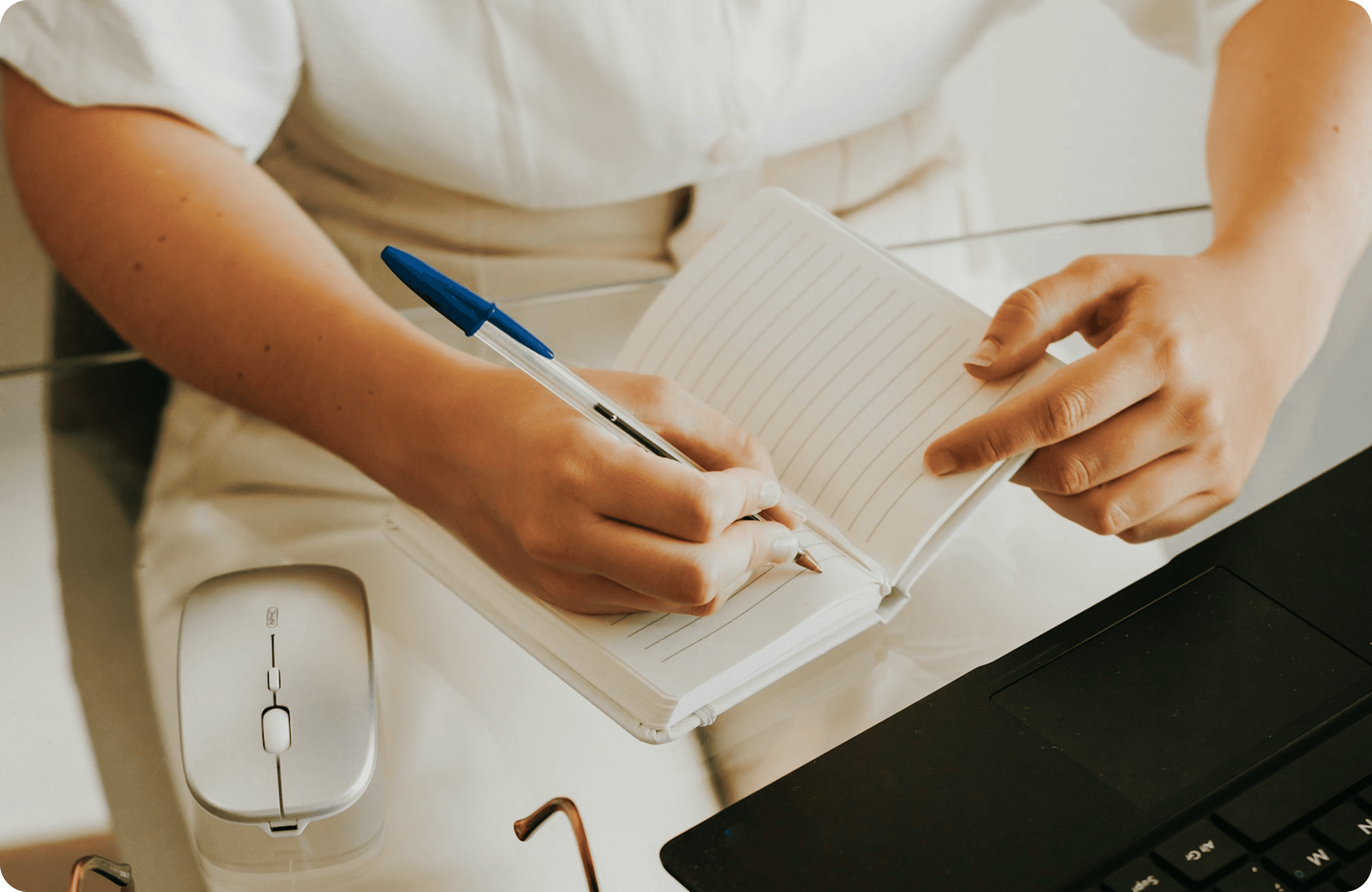 A person is writing in a lined notebook with a blue and white pen while sitting at a glass desk with a wireless mouse and a laptop nearby.