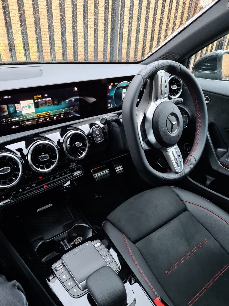  A vertical shot from the back seat of a car showing a woman driving. She is wearing a brown jacket, and her right hand is raised toward the open sunroof. The steering wheel has a Jeep logo. The road and trees are visible through the windshield.