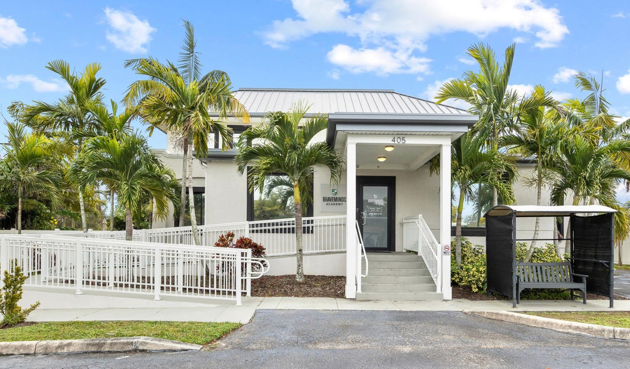Small white building with a ramp and steps, surrounded by palm trees under a clear blue sky. A bench sits near the entrance, conveying a welcoming feel.
