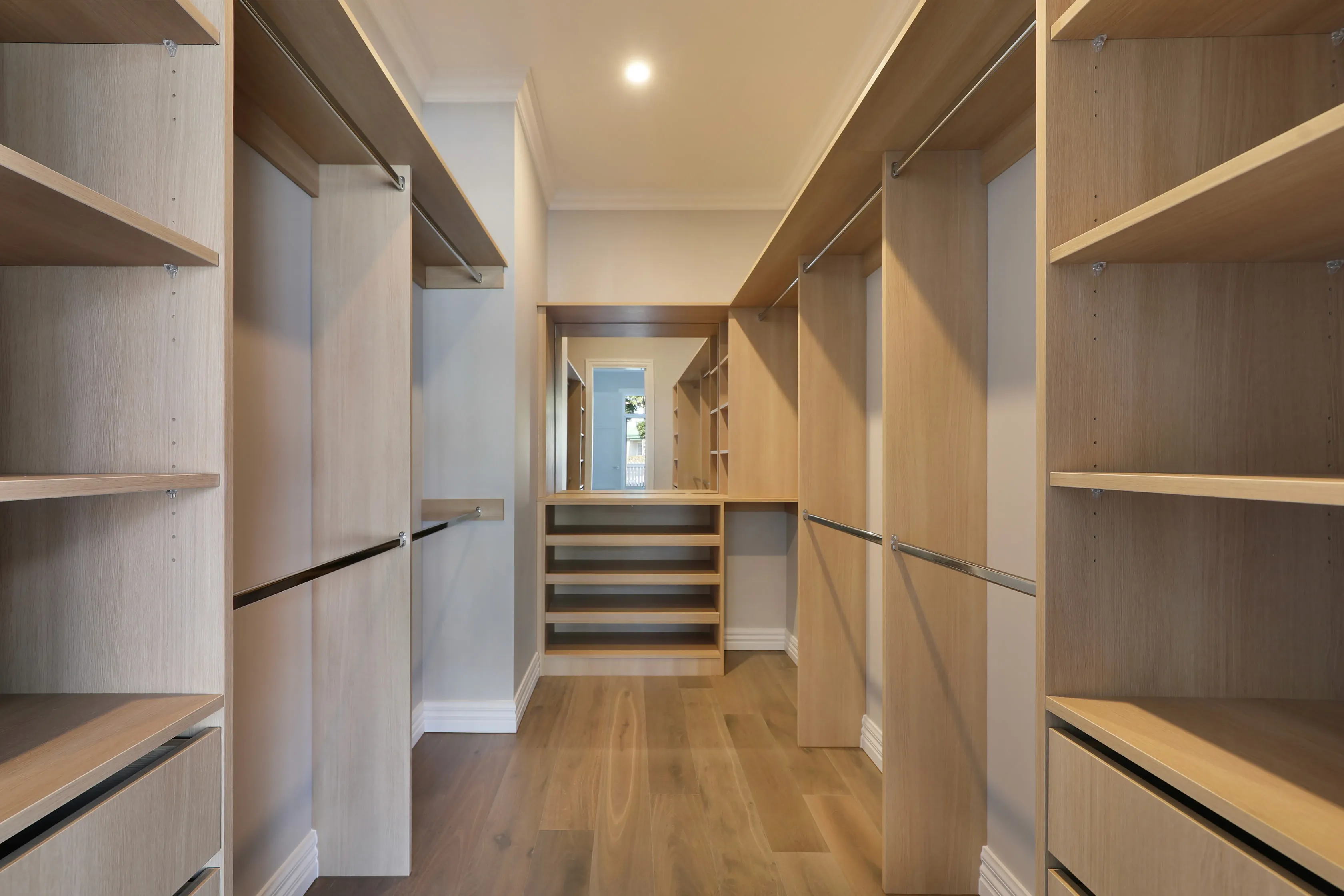 Walk-in robe with pale-oak floor-to-ceiling joinery, open shelving and hanging rails on both sides, a central bank of drawers below a mirror and oak floors