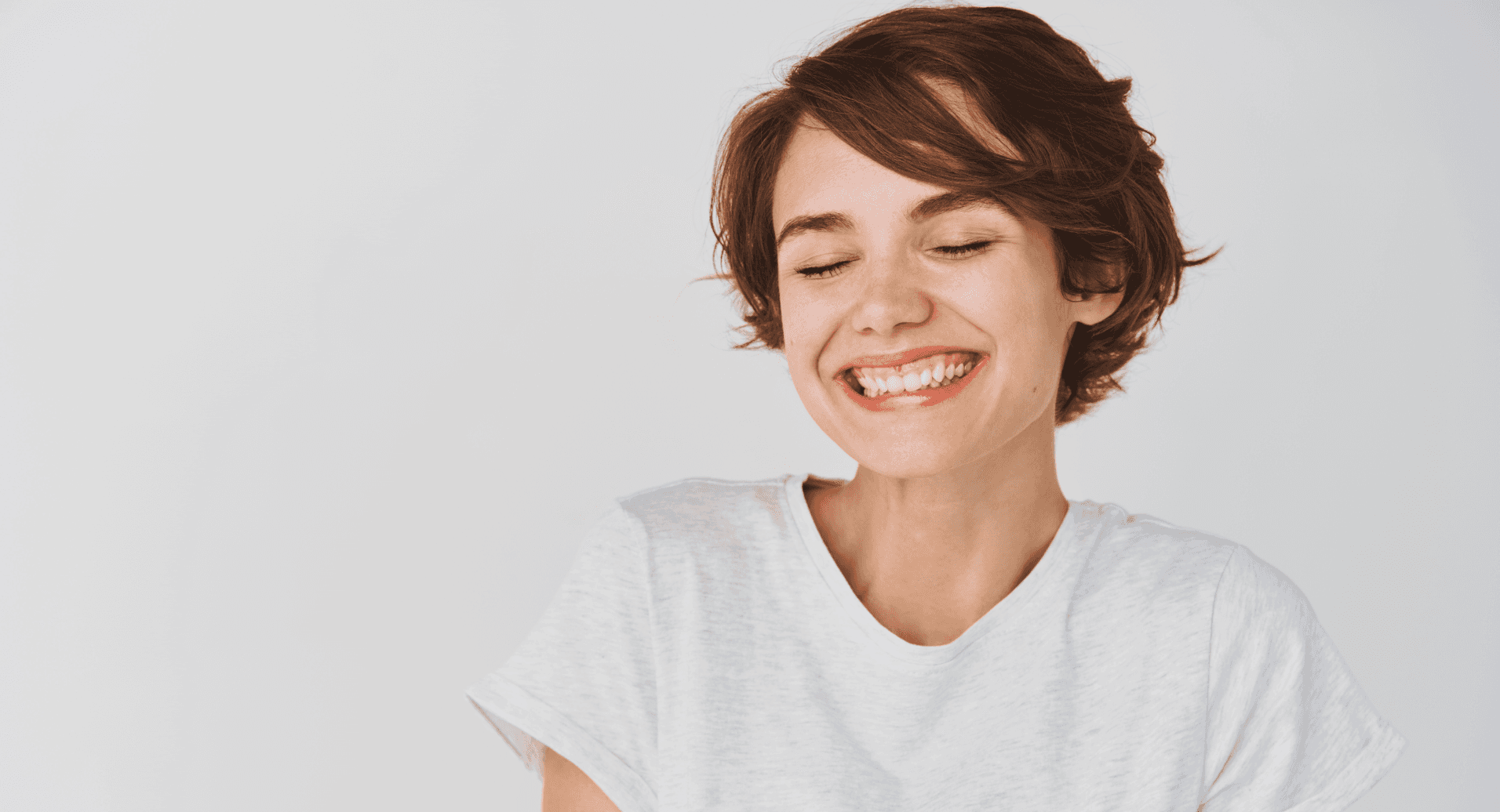 A young woman with short hair, wearing a white shirt, smiling with her eyes closed, looking happy and content