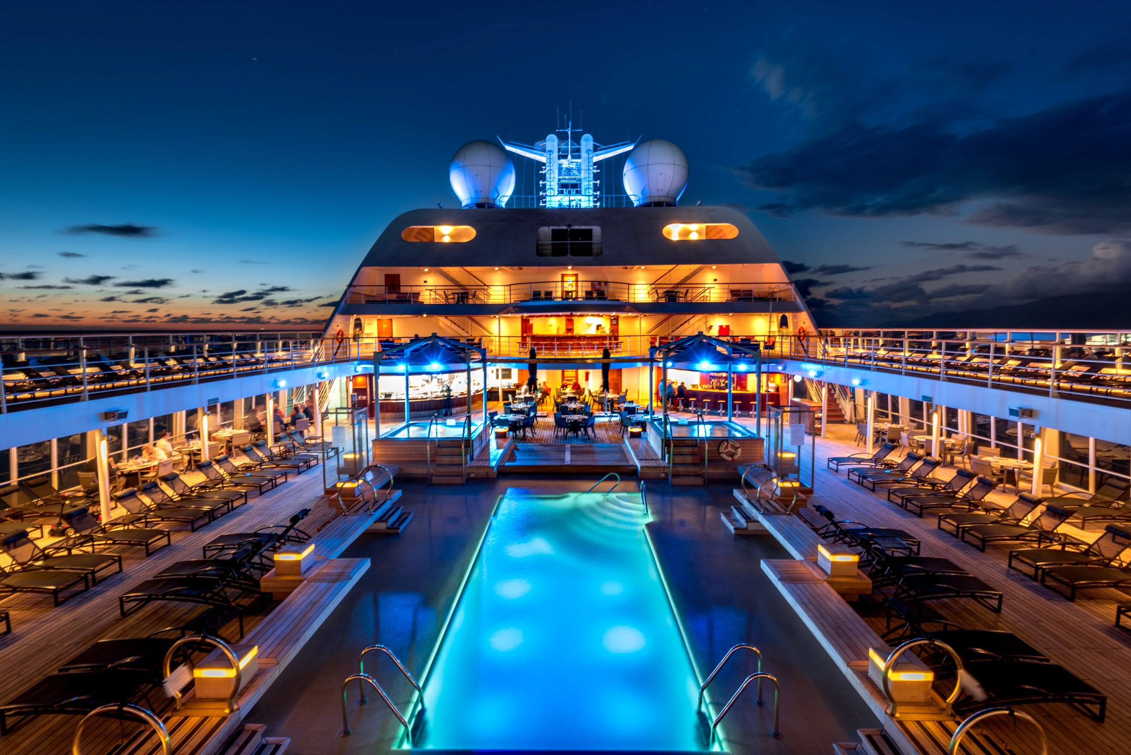 A large cruise ship features an illuminated outdoor pool deck with lounge chairs and a glowing blue swimming pool at night.