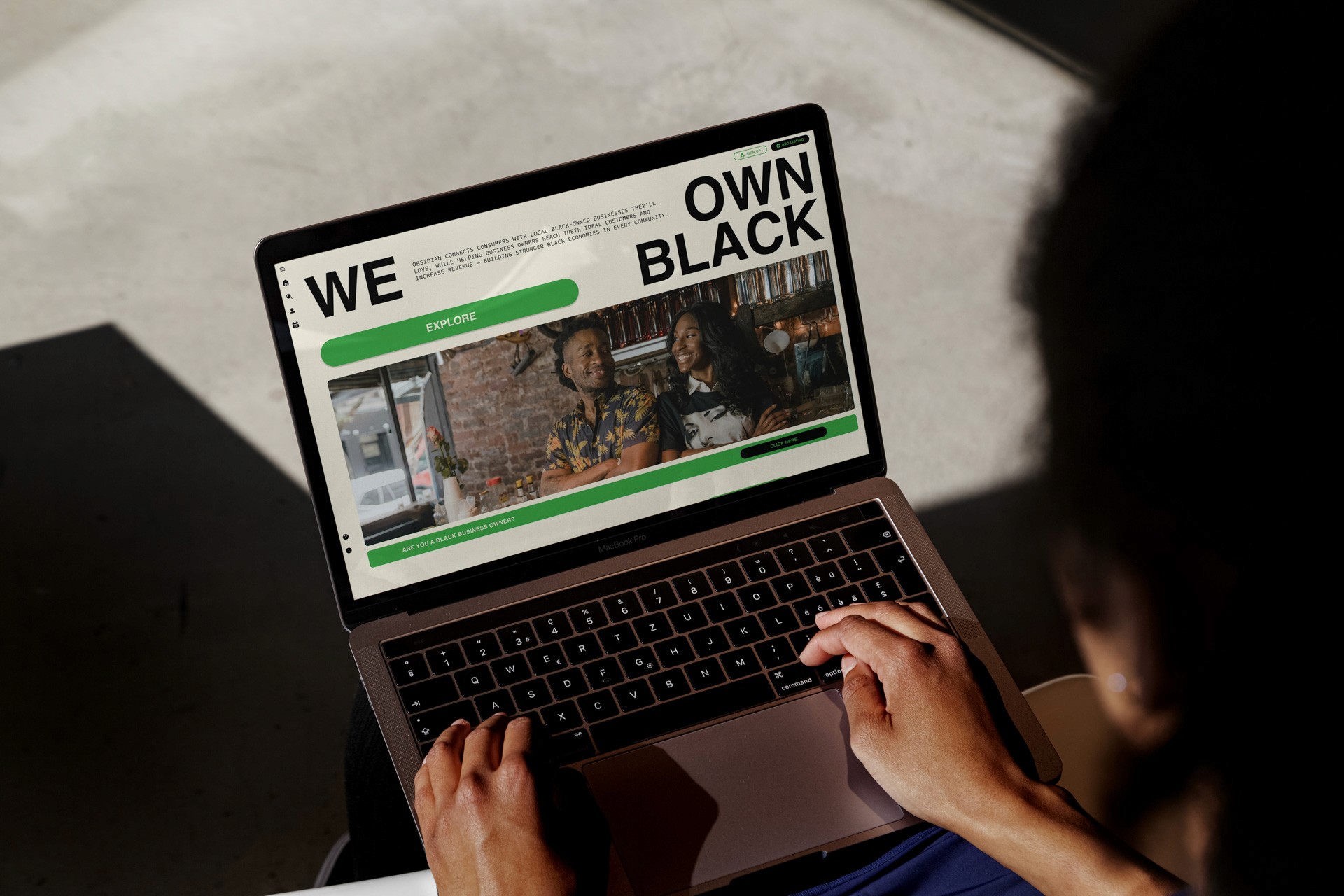 A person with dark hair is using a laptop. The laptop screen displays a webpage with the title "WE OWN BLACK" in large text at the top. Below the title, a photograph shows two women, one of whom is wearing a patterned top. The webpage also has a green "EXPLORE" button. The laptop is resting on a light-colored surface, and the person's hands are positioned on the keyboard.