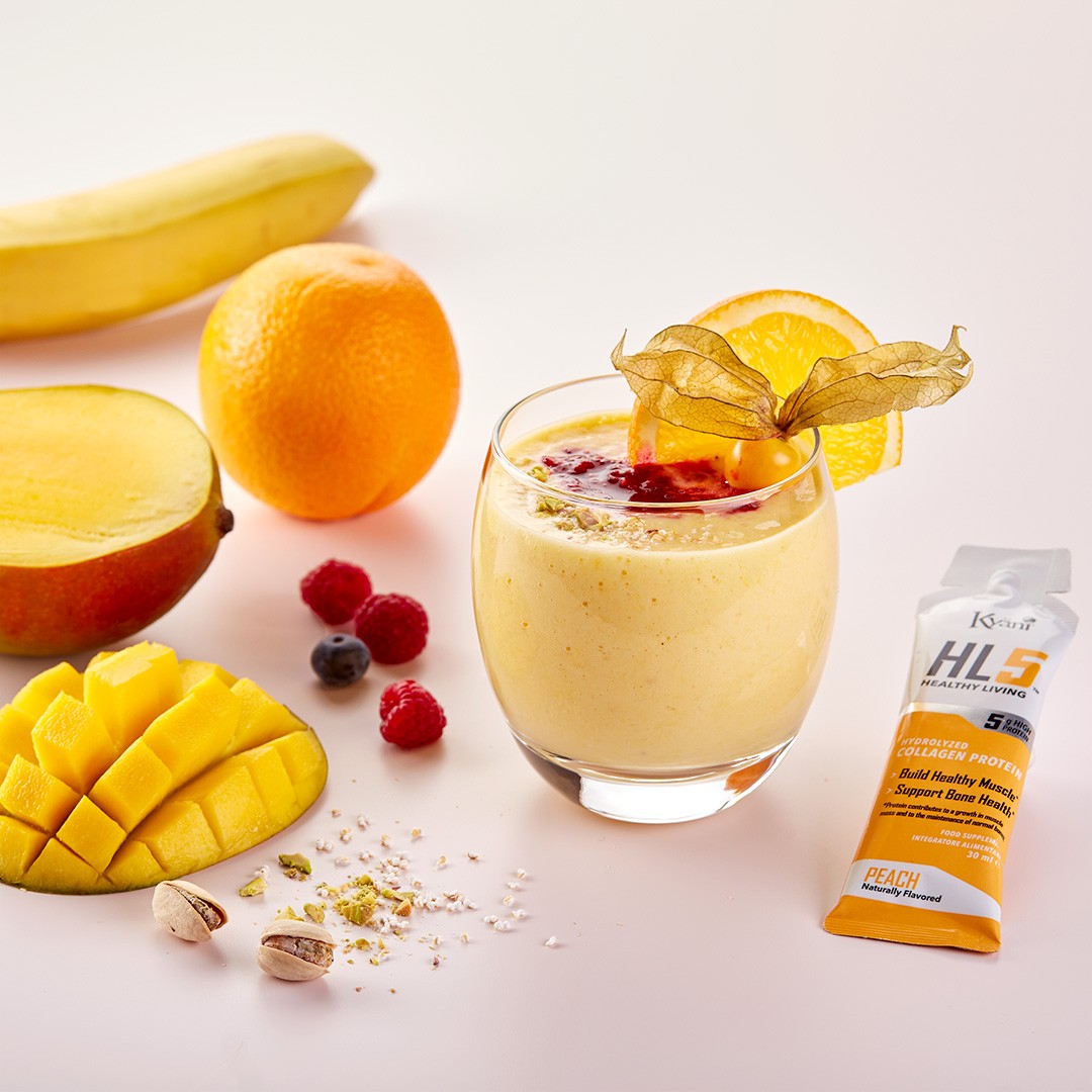 A refreshing Kyäni smoothie is elegantly displayed in a clear glass, garnished with an orange slice and gooseberry, surrounded by fresh fruit like mango, banana, and orange, along with a sachet of Kyäni HL5 Peach protein, on a clean, white background.