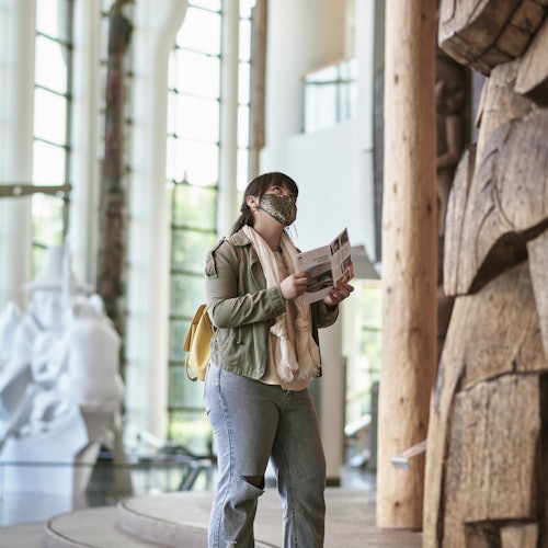 在一个宽敞明亮的博物馆里，一位戴着口罩、手持宣传册的人站在一个大型木雕附近。