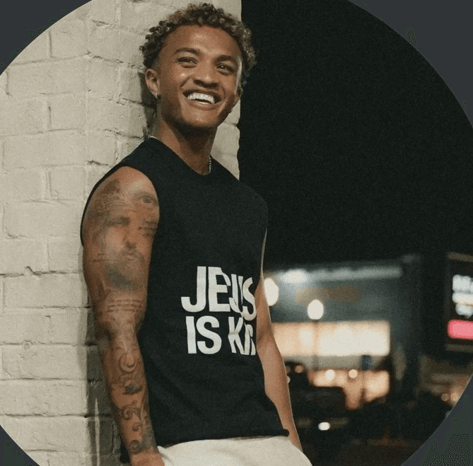 Smiling man with windswept red hair outdoors, wearing a black jacket and green backpack. Bright sunlight and a blurred natural background convey joy and adventure.