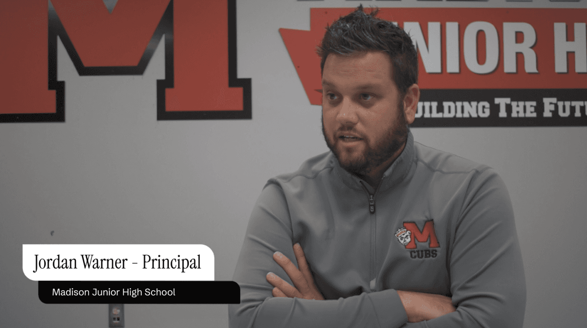 A young woman with long dark hair stands outdoors at night beside a blue vehicle. She is wearing a black athletic-style jersey with white stripes and graphic elements on the front. The background shows softly lit buildings and warm lights, suggesting an urban setting. She appears to be mid-step, looking slightly to the side with a composed expression.