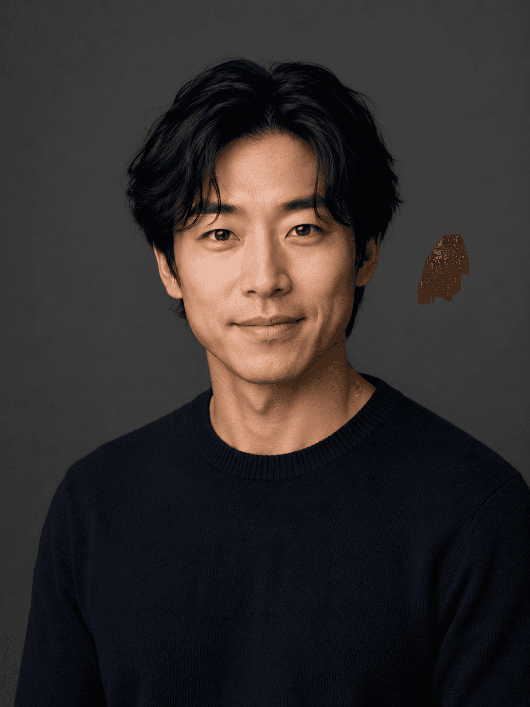 A young man with dark hair, wearing a black shirt, smiles softly against a plain, dark background.