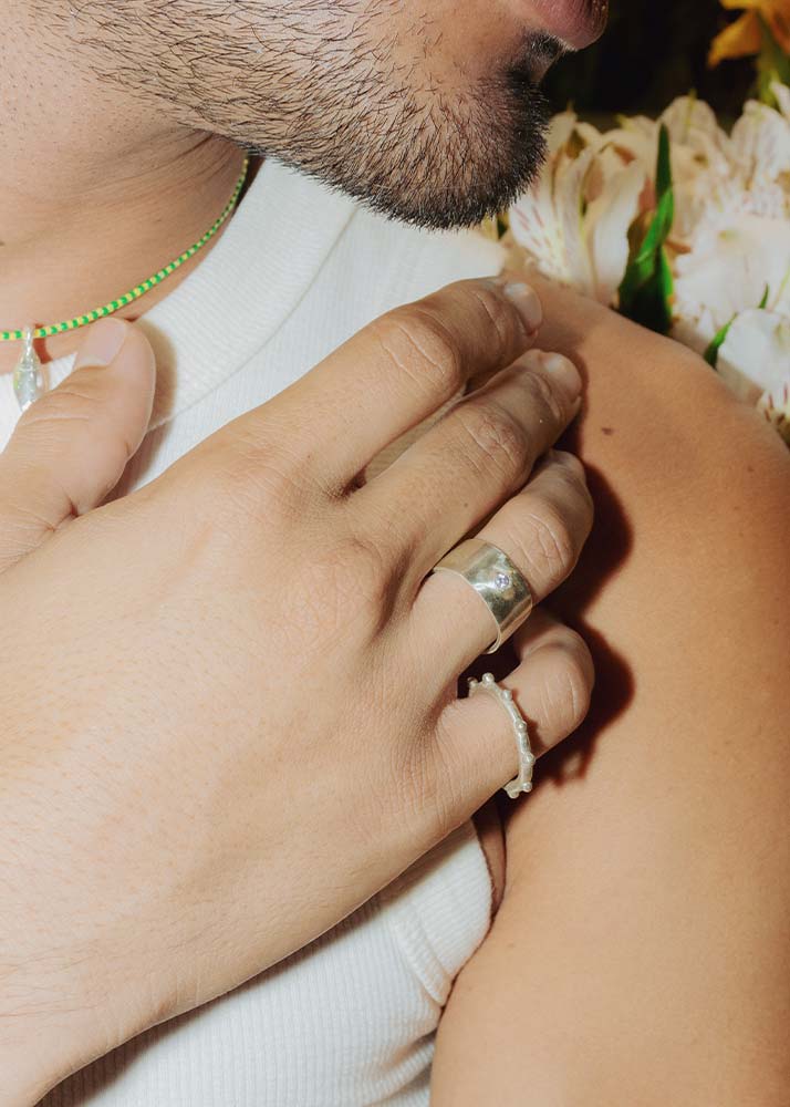 Close-up of a person's hand resting on their shoulder, wearing a wide silver band ring with a clear central gemstone and a smaller textured silver ring.