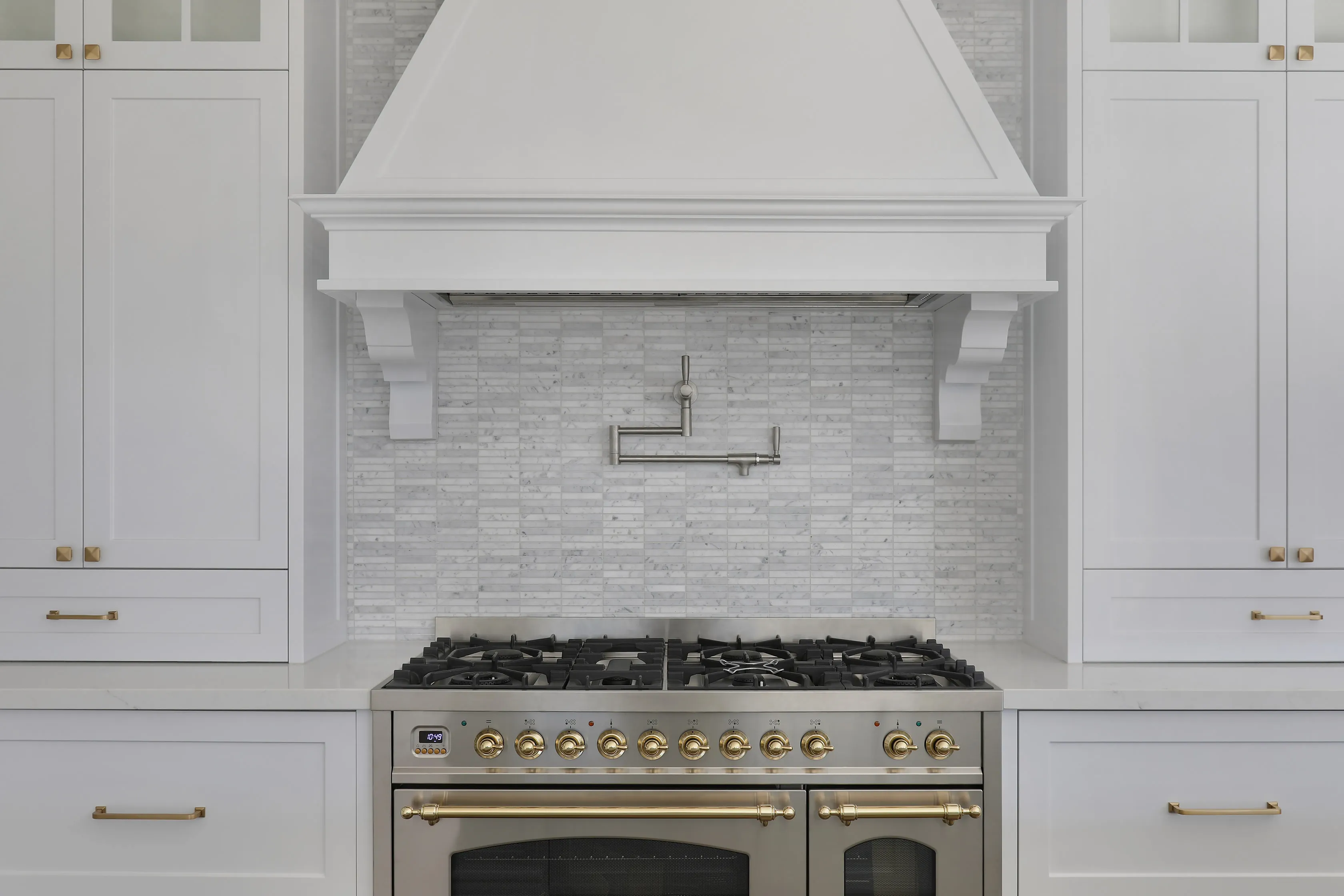 Kitchen detail of a white mantle-style range hood over a stainless and brass-knobbed gas range, with a brushed-nickel pot filler and a marble mosaic splashback flanked by white shaker cabinets