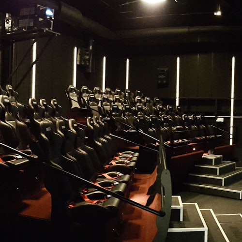 Hämärästi valaistu teatteri, jossa on useita rivejä mustia ja punaisia liikemahdollisuudella varustettuja istuimia, jotka ovat suunnattu projektoria ja näyttöaluetta kohti.