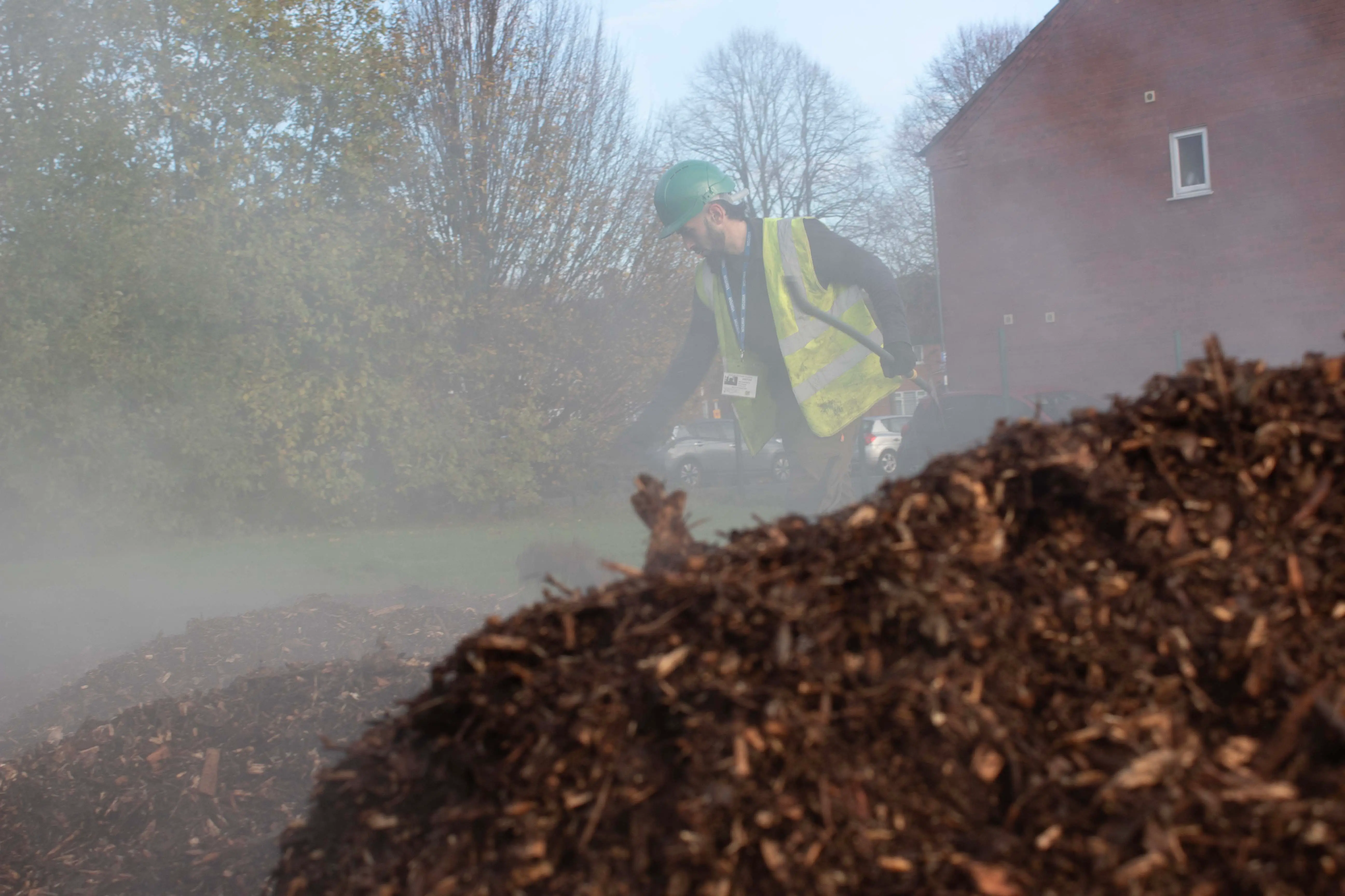 A construction site with a large pile of soil or mulch, partially obscured by mist and trees in the background.