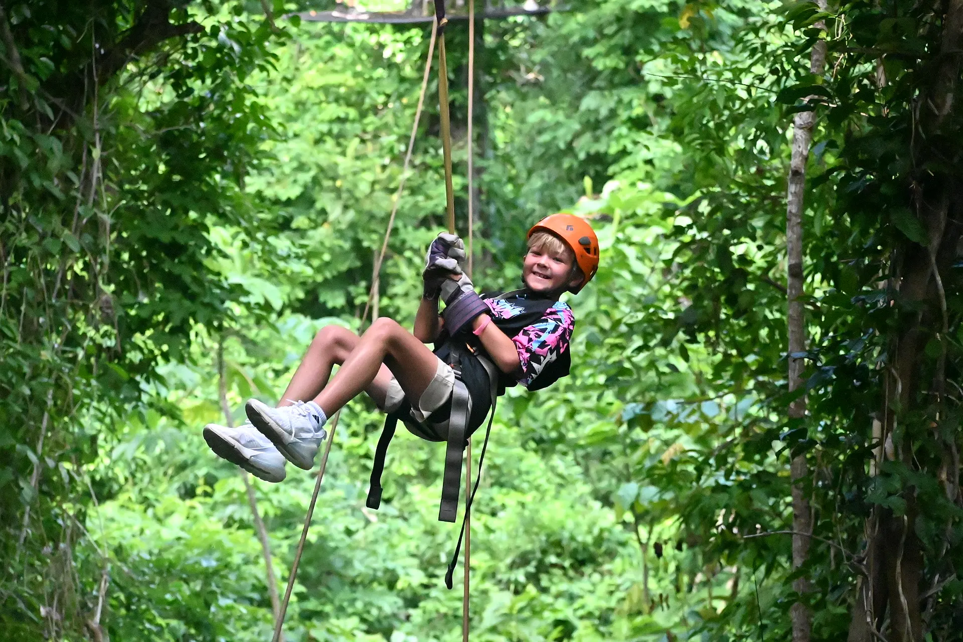 Smiling child in a helmet and harness riding a zipline through green forest canopy