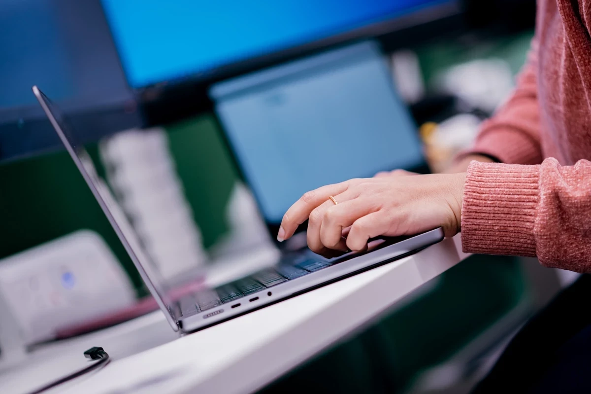 Close-up of hands typing on a laptop placed on a white desk, with another open laptop and monitor in the background.