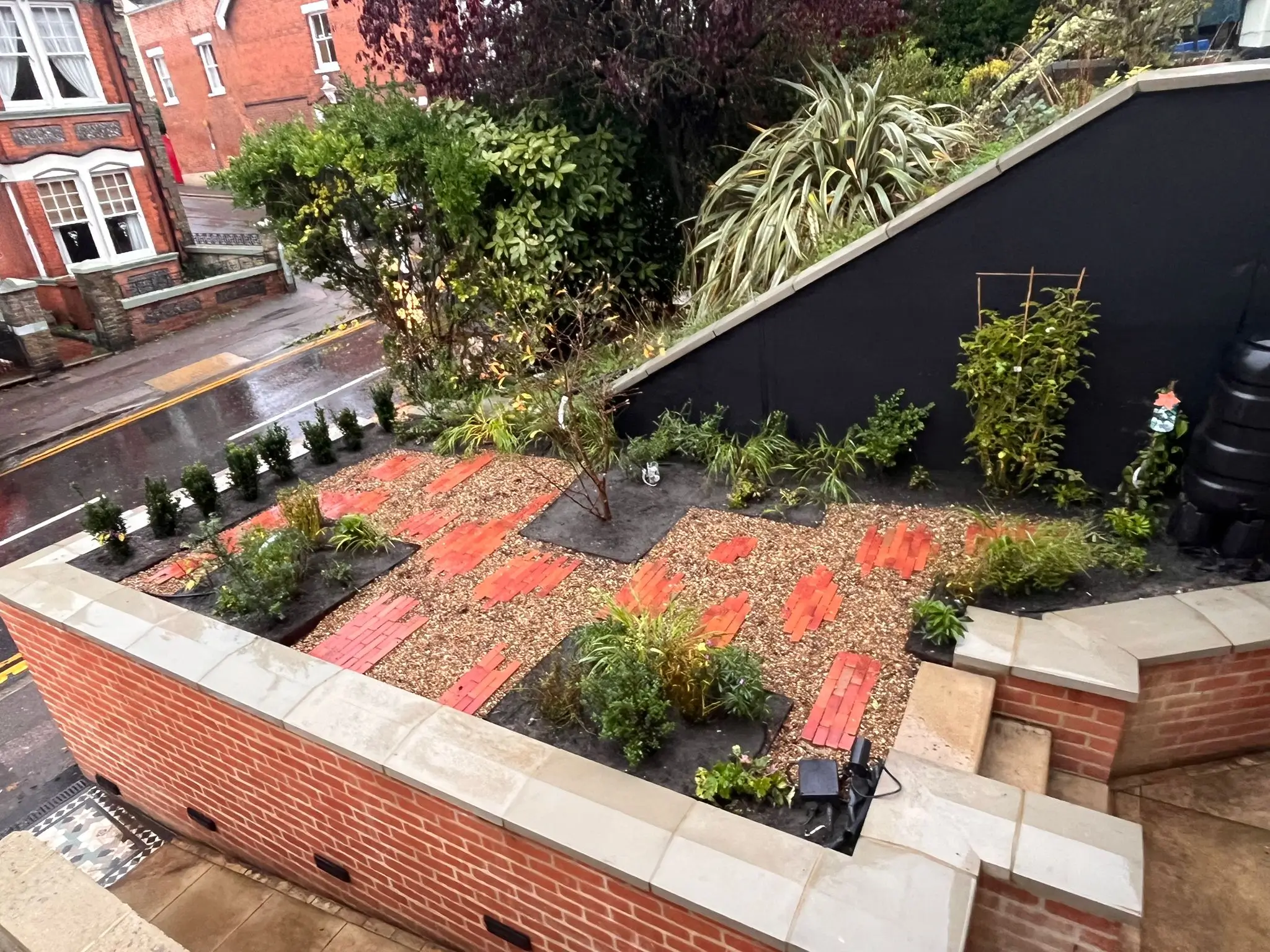 A landscaped garden featuring brick paths, greenery, and steps, set on a slightly sloped area beside a building.