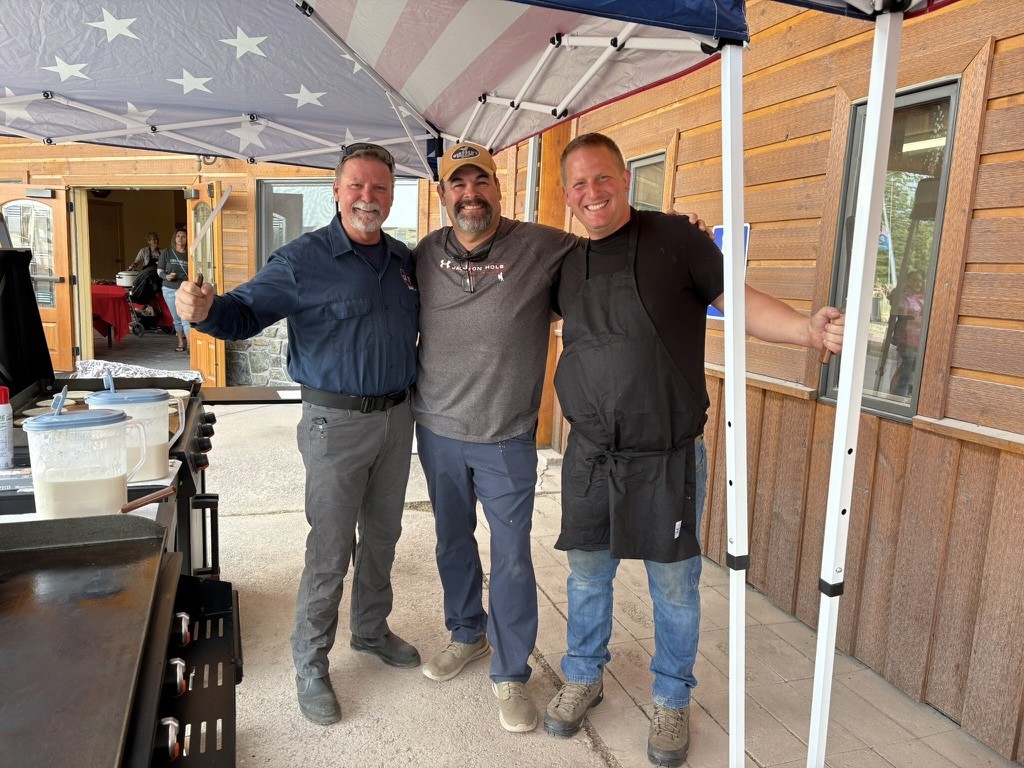 Three men standing together beneath a canopy beside a serving table.