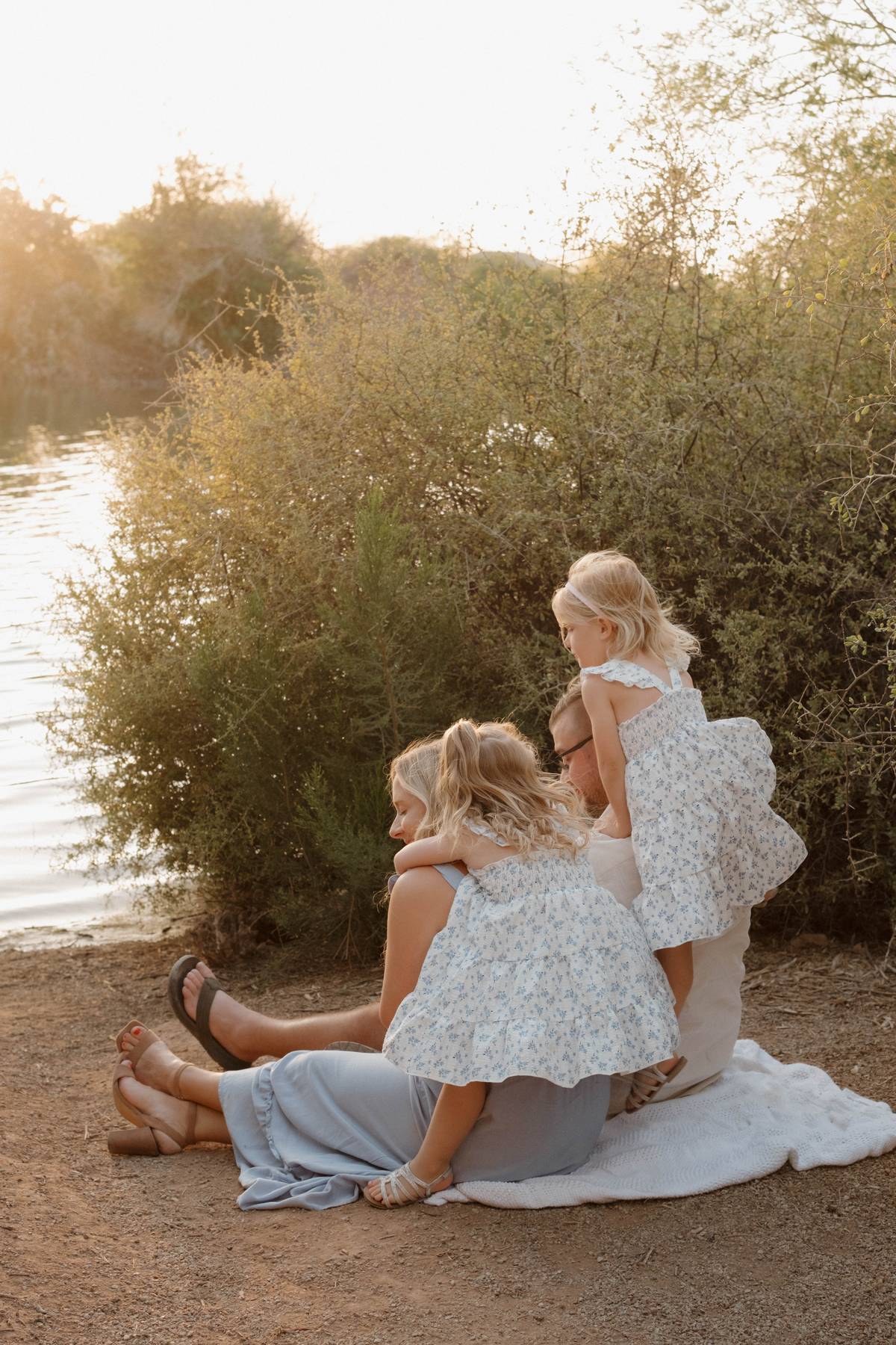 Family of four sitting by a lake at sunset, with two young girls in white dresses looking at the water, and parents with a baby in the foreground during a lifestyle family photoshoot, in Gilbert, Arizona.