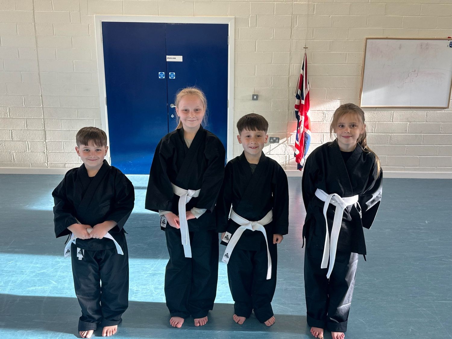 Four children in black martial arts uniforms with white belts stand smiling in a hall.