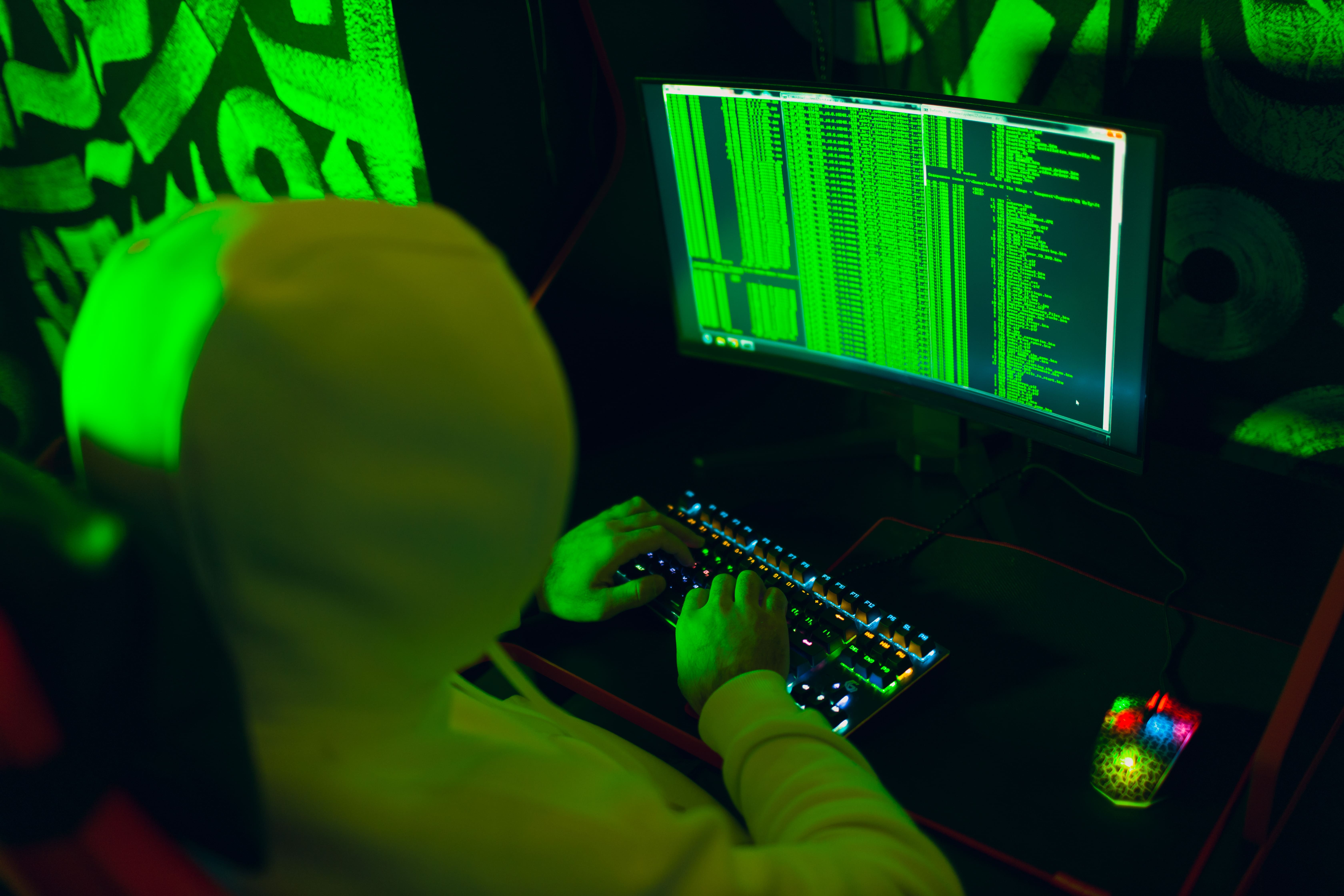 Hacker in hoodie using computer with green code on screen in dark room, symbolizing cybersecurity breach or data theft
