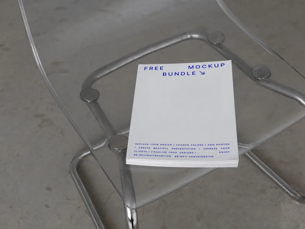 A stack of booklets with a clean white cover placed on a transparent acrylic chair. The design features minimal blue text and is set against a neutral concrete floor with soft lighting.