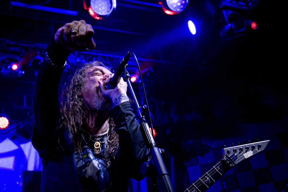 Energetic shot of a metal rock vocalist singing passionately into a microphone on a stage lit with intense blue spotlights.