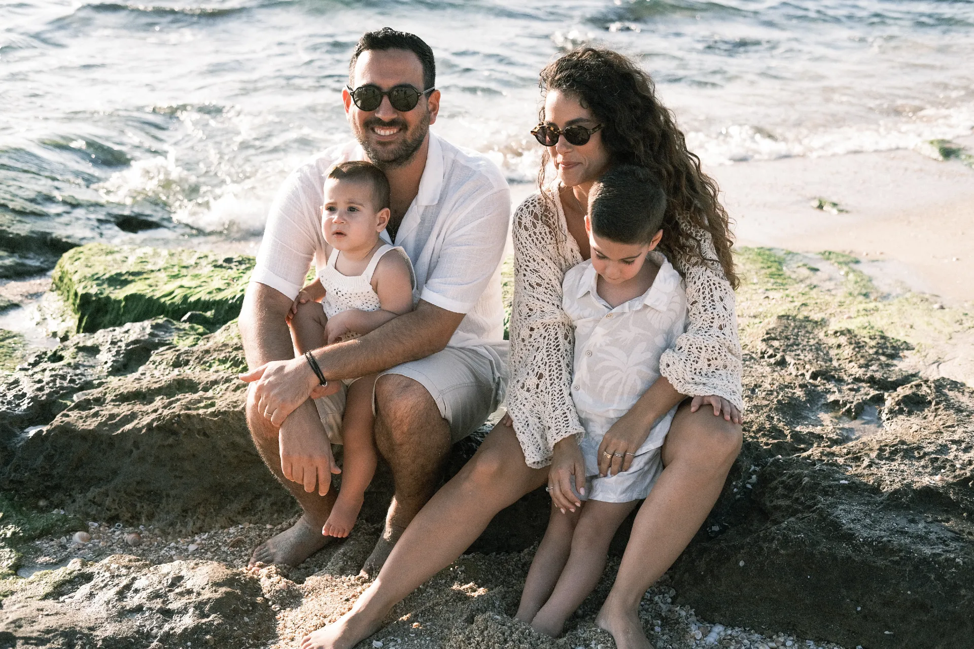 צילום דיוקן משפחתי על חוף הים בשעת בין ערביים. ההורים ושני ילדיהם יושבים על סלעי החוף בלבוש לבן וקליל, באווירה קיצית וטבעית מול הגלים, מאת Shiriz Photography והצלמת שירי עציץ