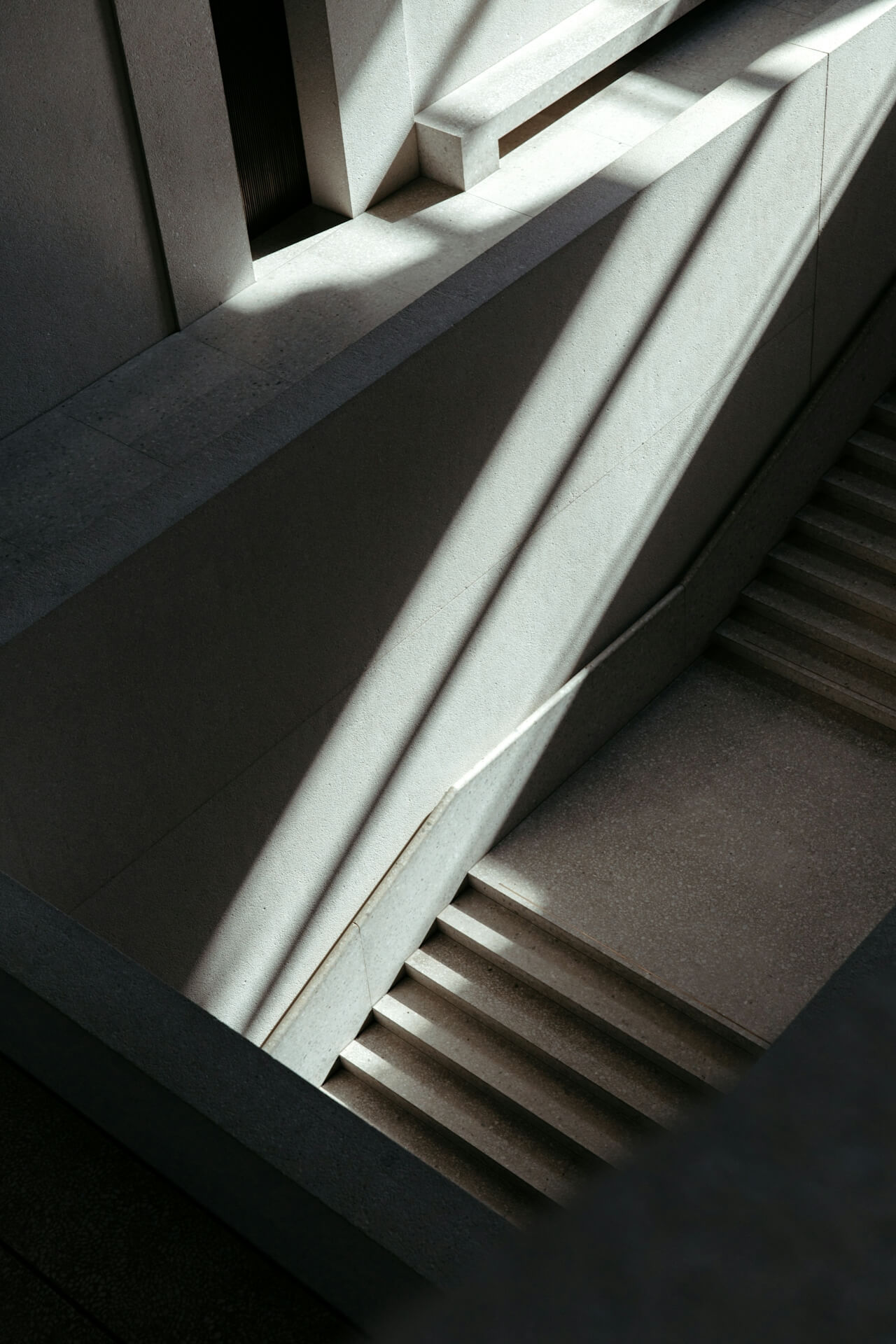 Sunlight casts geometric shadows on a minimalist stairwell with concrete steps and walls, creating a serene and contemplative atmosphere.