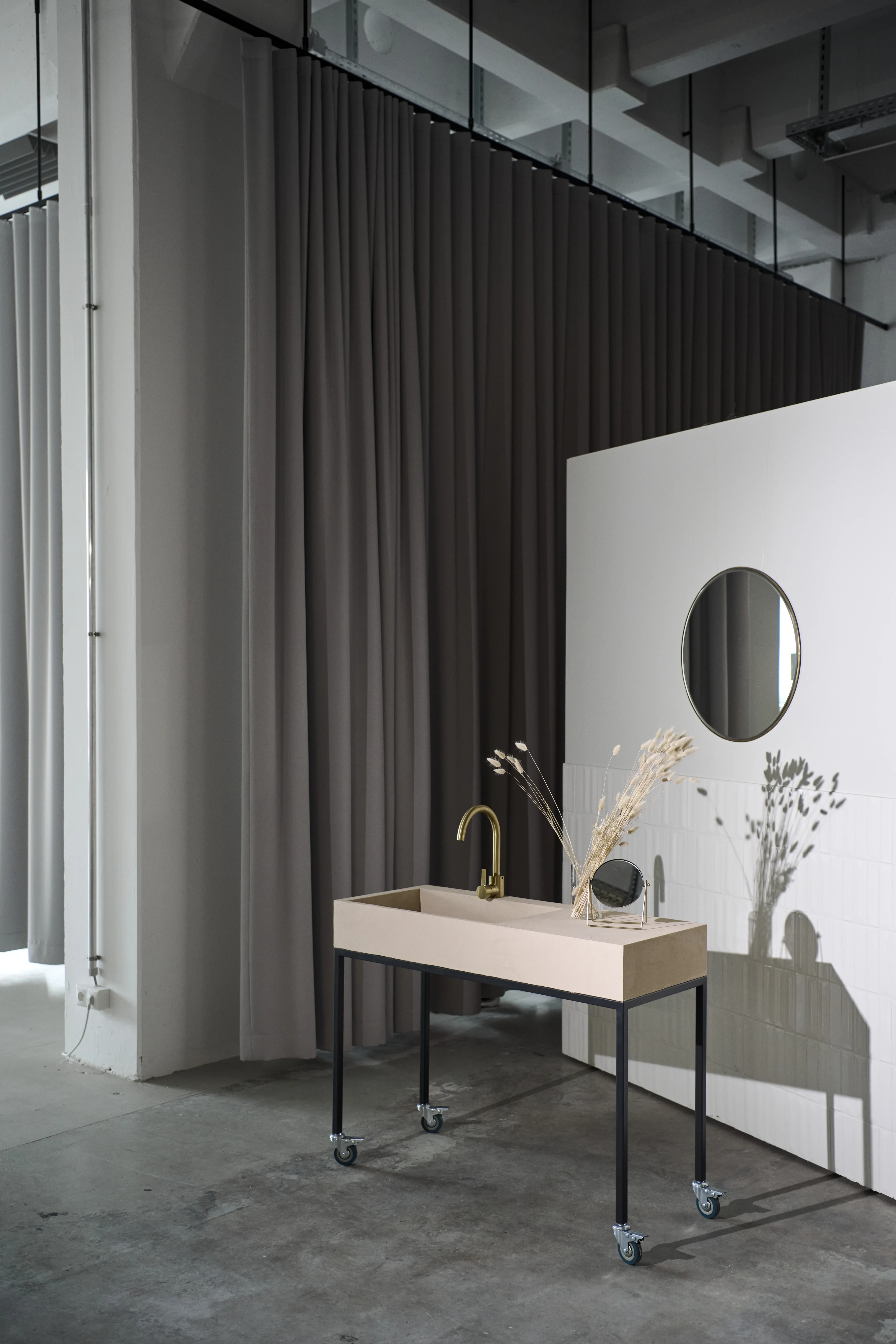 Mobile washbasin unit at the FLACONI CONTENT STUDIO with a beige stone trough, brass tap and black frame on castors, in front of a white tiled wall with round mirror.