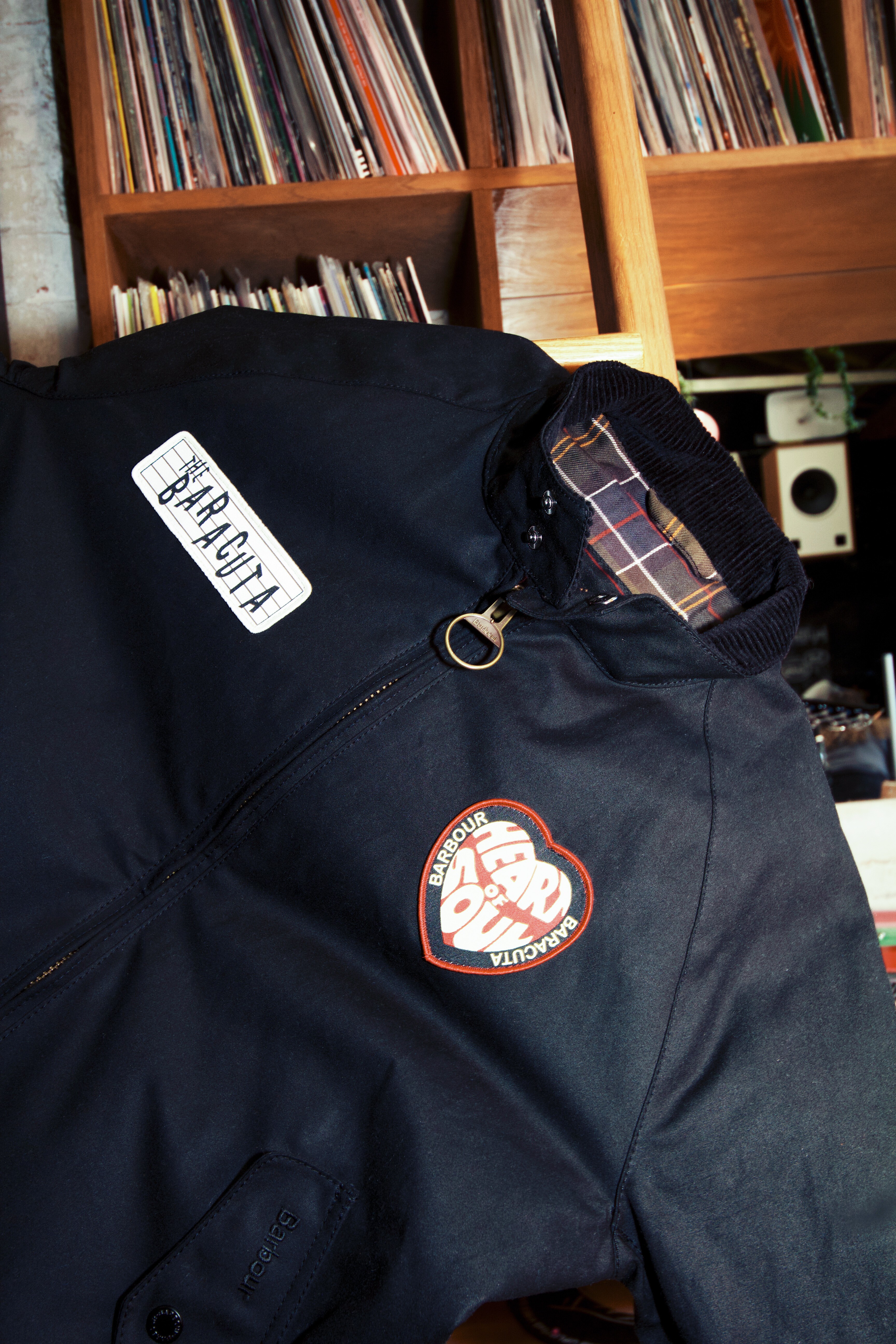 Détail de blouson Baracuta x Barbour, cotonbleu marine, doublure tartan et patches, devant un mur de disques vinyl