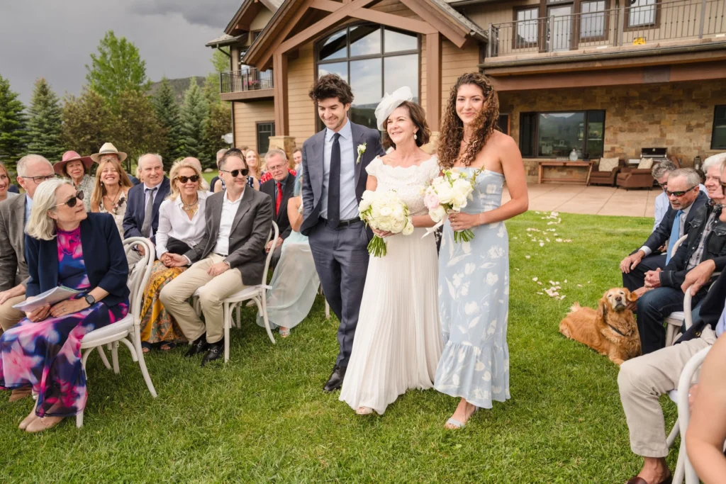 A bride in a white gown and a bridesmaid in a light blue patterned dress walk across a green lawn past seated guests at a Chaparral Ranch Wedding.