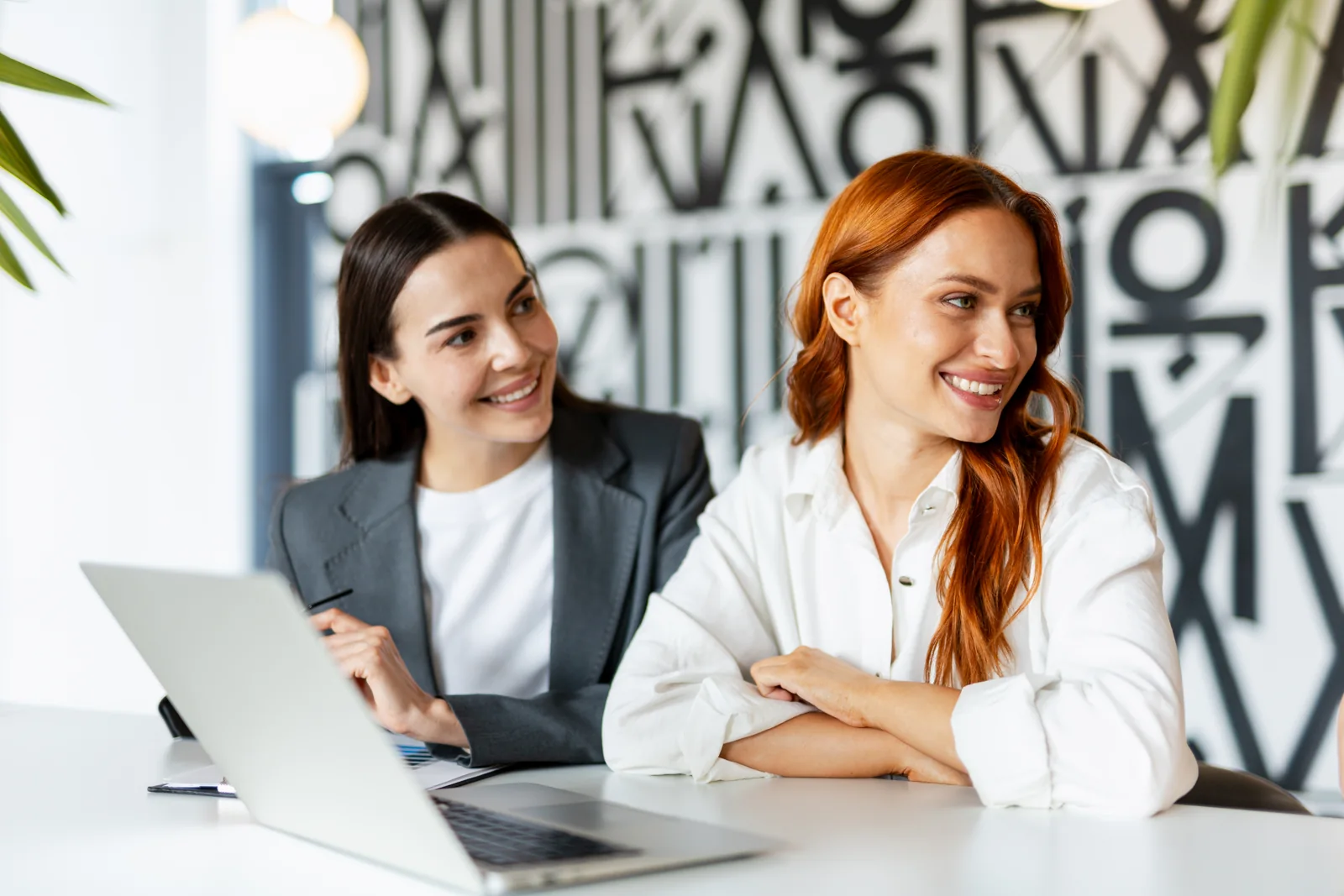Two businesswomen smiling together at a desk with a laptop