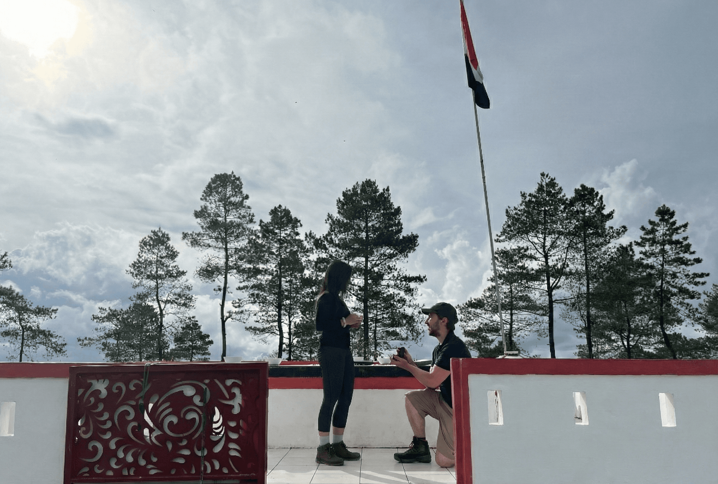 A couple holds hands, standing on a rocky beach under a large cave arch, gazing at the tranquil ocean during sunset. The sky is softly lit in pastel hues.